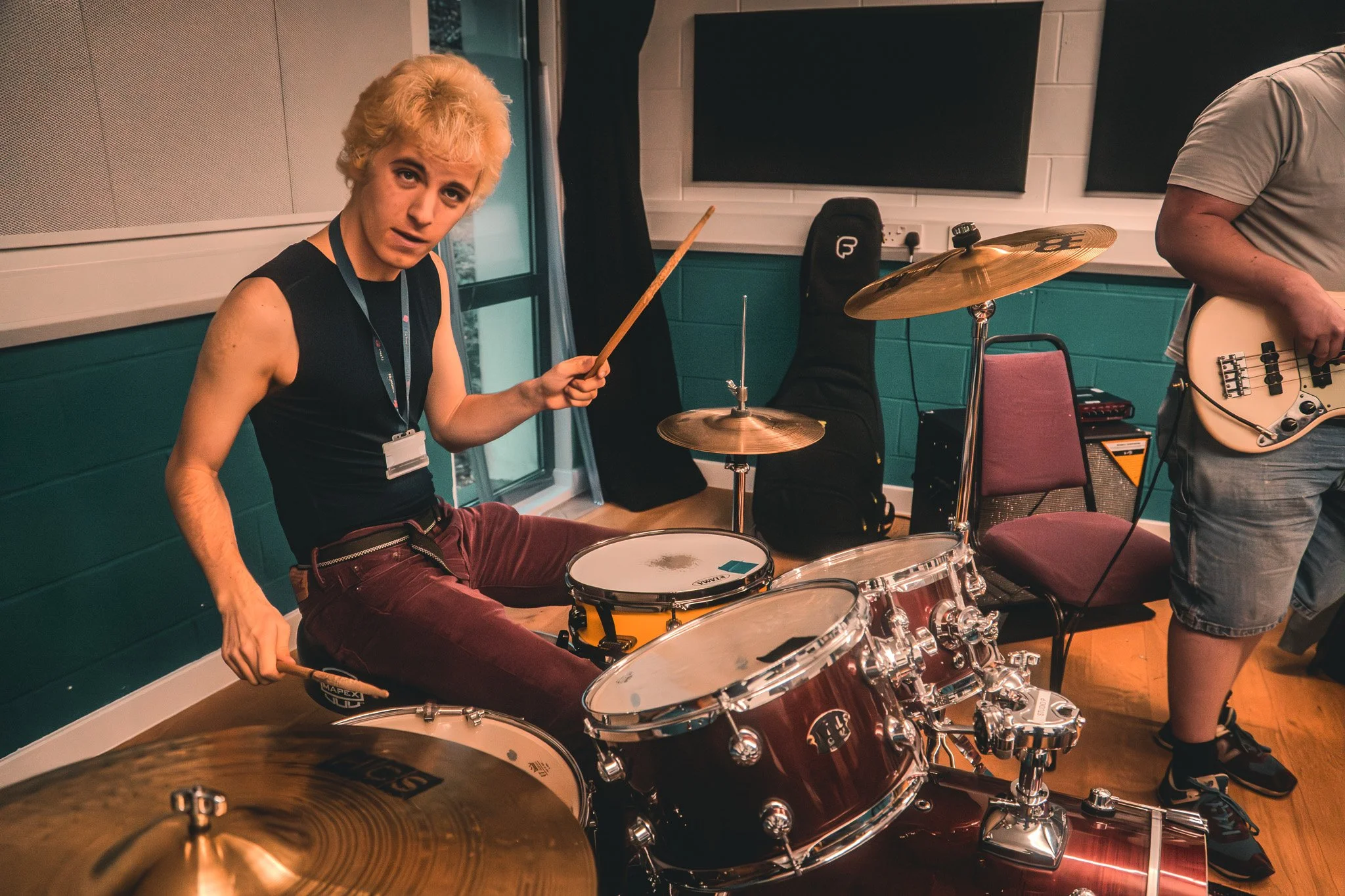 A young man with blonde hair sitting on a drum kit, holding drumsticks, looking at the camera. He's in a music rehearsal room with teal walls, there’s another person partially visible with a bass guitar, and various musical equipment in the backgroun