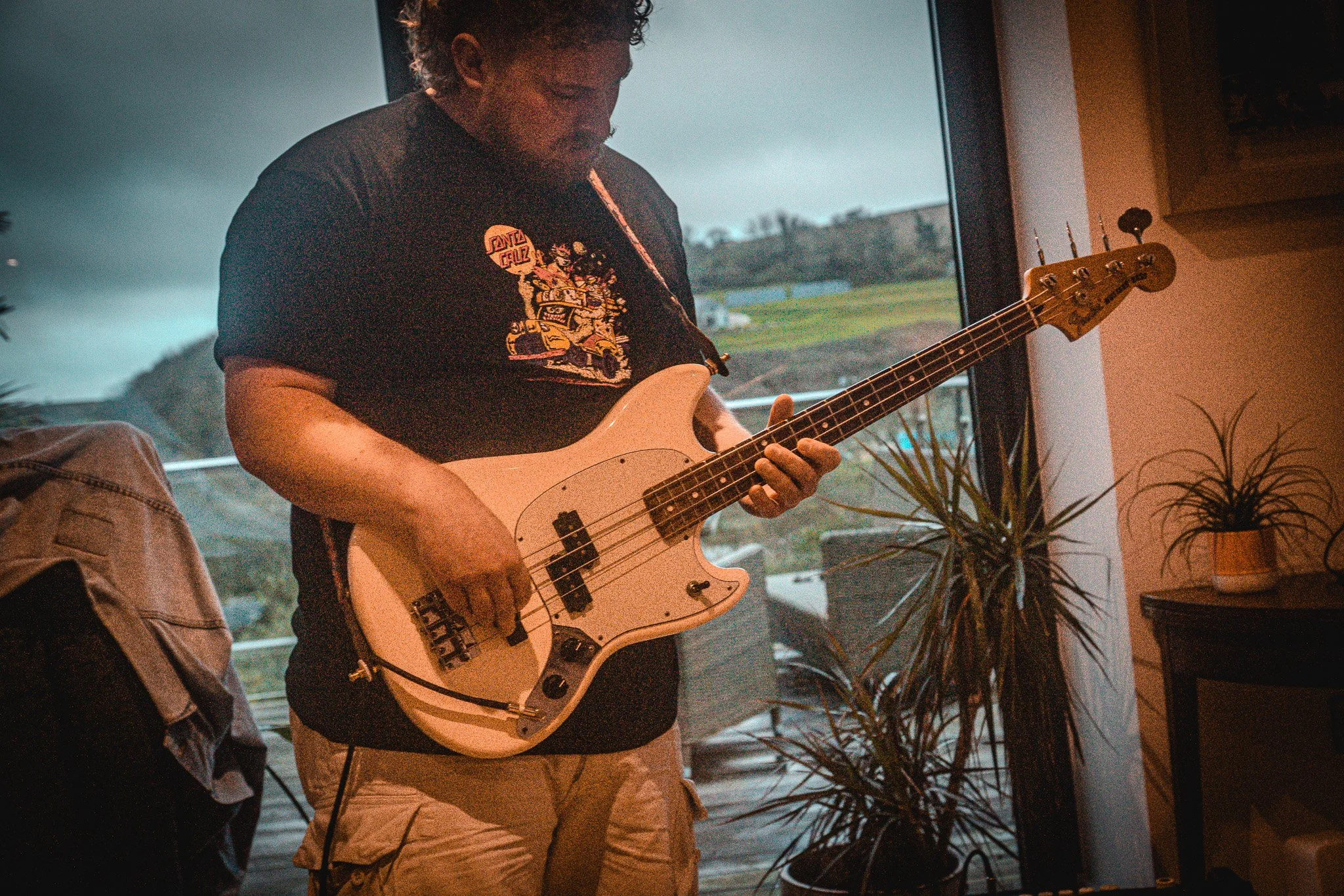 A man playing a white electric bass guitar indoors, with plants and large window showing an outdoor landscape in the background.