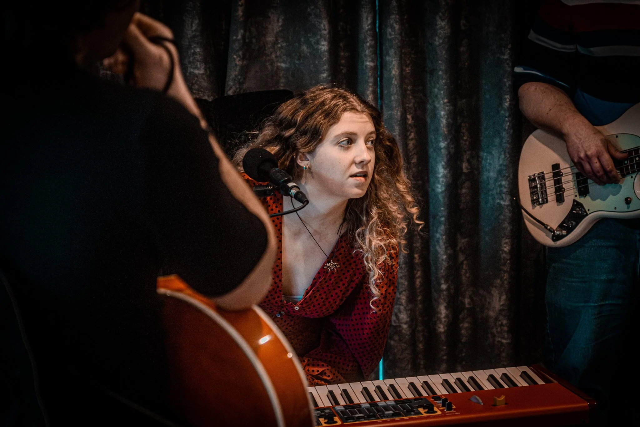 Young woman with curly hair playing a keyboard, surrounded by musicians with guitars, in a dimly lit setting with dark curtains.
