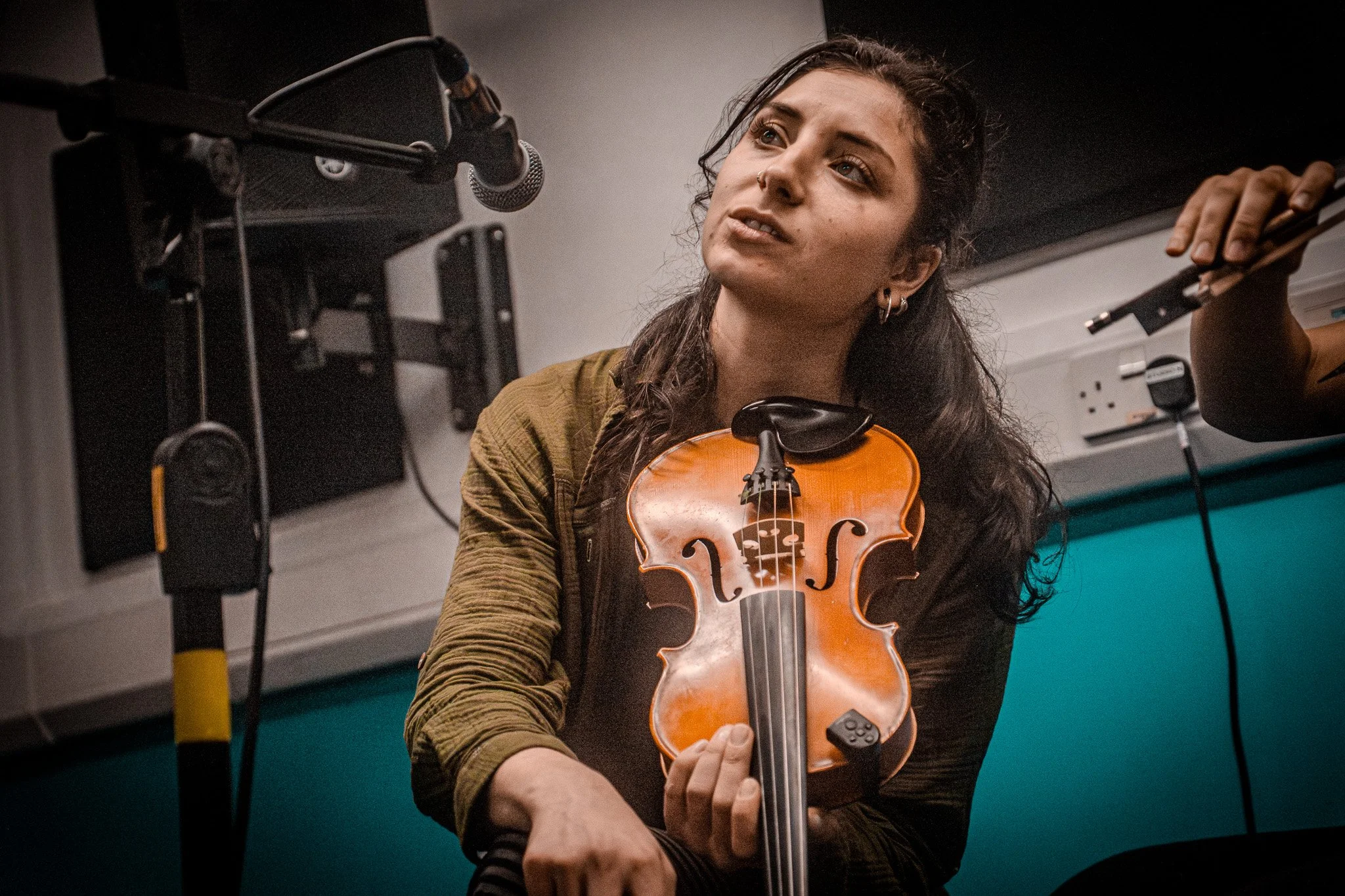 A woman with long dark hair holding a violin and looking thoughtfully while sitting near a microphone in a music studio.