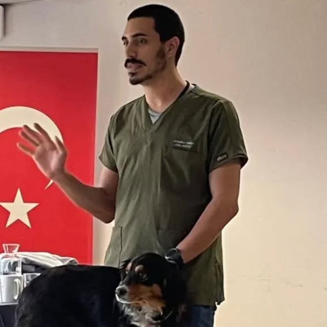A man in medical scrubs speaking, with a black and brown dog sitting on a table in front of him. A red flag with a white star and crescent moon is in the background.