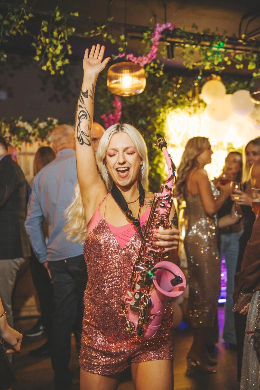 A woman in a pink sequin dress playing a pink saxophone at a party or celebration, with people dancing and socializing in the background.