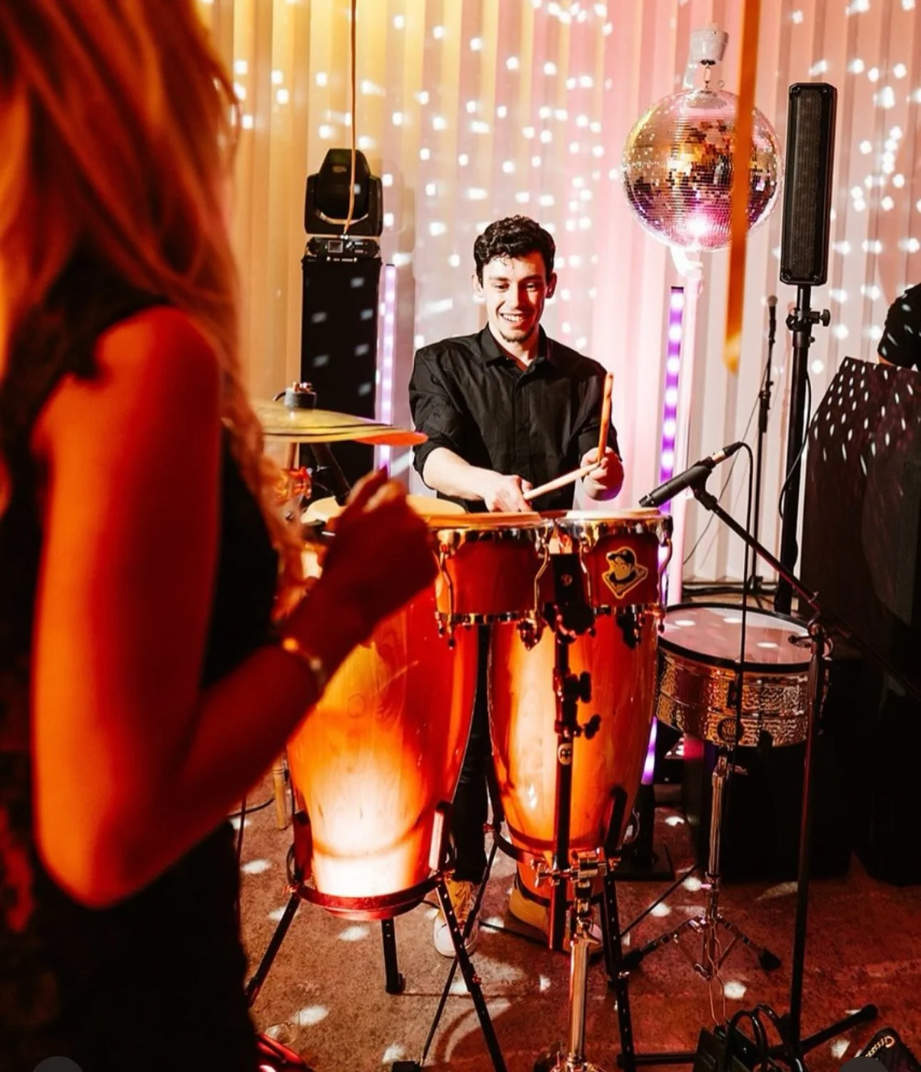 A man playing bongos at a party with a woman standing nearby. The room is decorated with colorful lights and a disco ball.