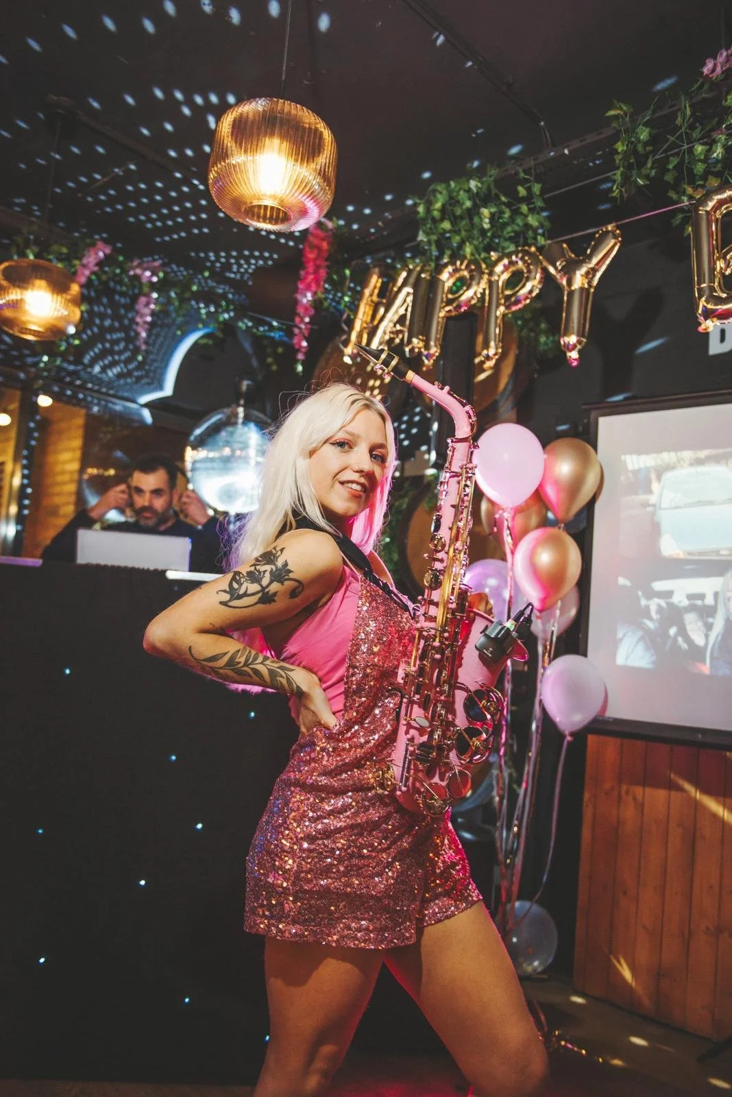 A woman with long blonde hair and tattoos on her arm, wearing a pink sequin dress, holding a pink saxophone, at a celebration with balloons and banners, with a DJ in the background and a sign spelling 'HAPPY'.