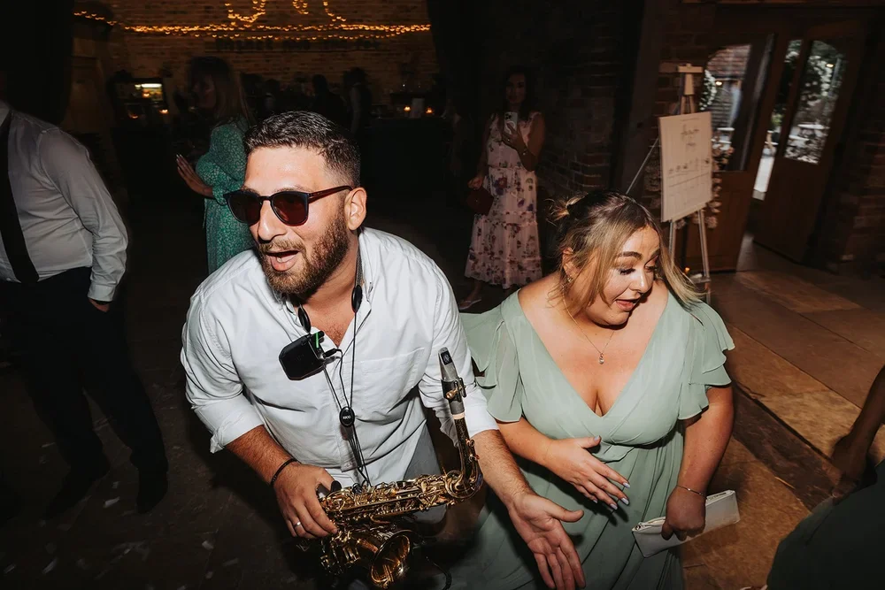 A man with sunglasses playing a saxophone at a party or event, with a woman in a green dress dancing and smiling nearby, in a warmly lit indoor venue.