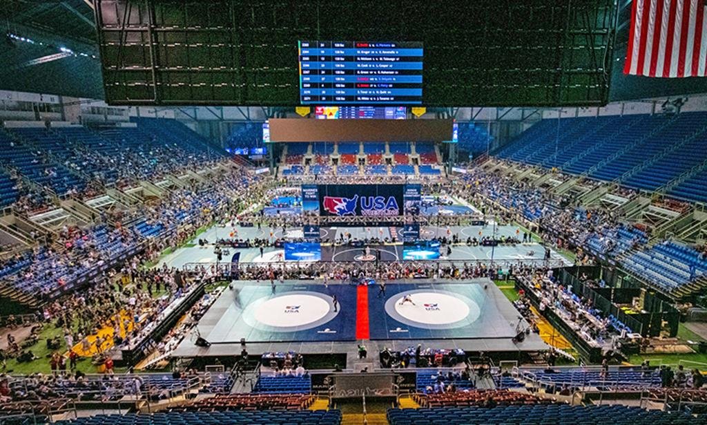 An indoor sports arena with a wrestling mat on the floor, surrounded by spectators and digital screens displaying USA and wrestling logos.
