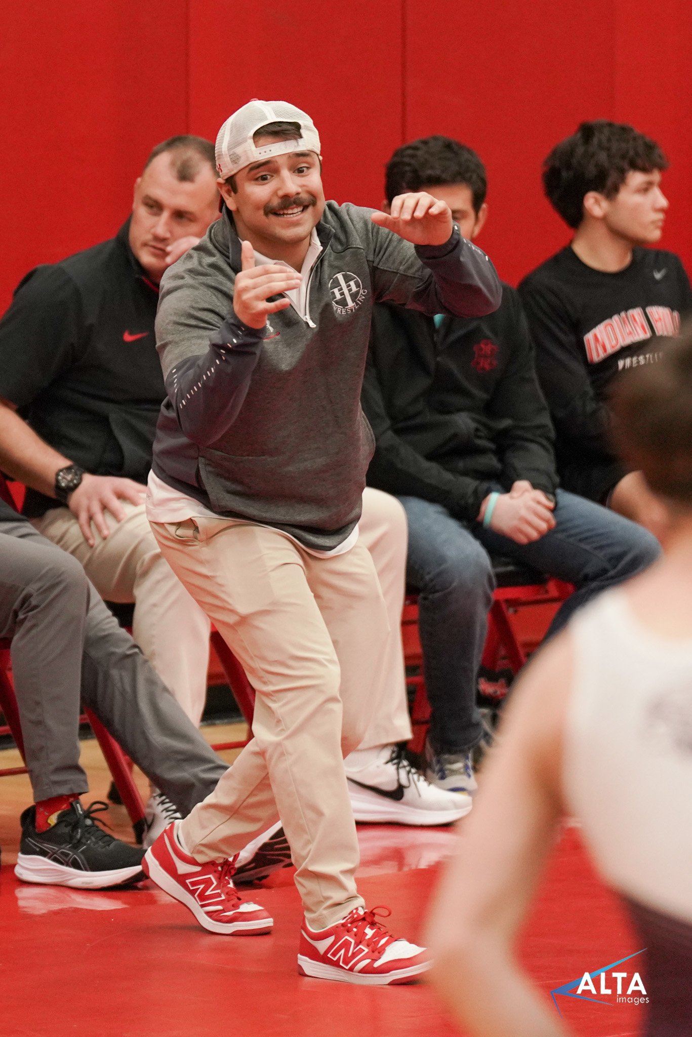 A coach or trainer in a gray jacket, beige pants, and red New Balance sneakers, standing and gesturing during an indoor wrestling event with wrestlers seated in the background.