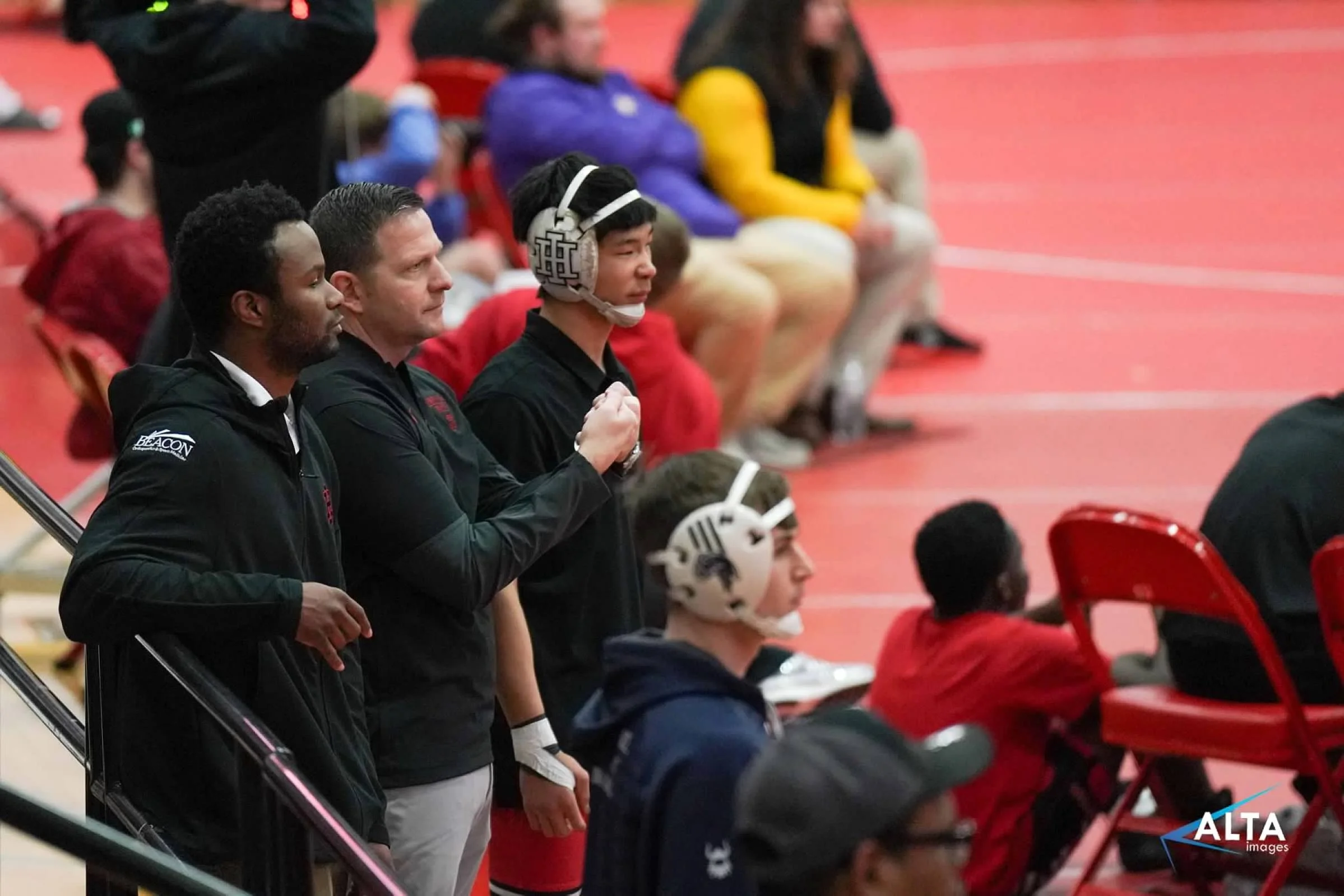 A group of athletes and coaches standing and sitting on the sidelines during a sporting event in a gymnasium, some wearing protective helmets.