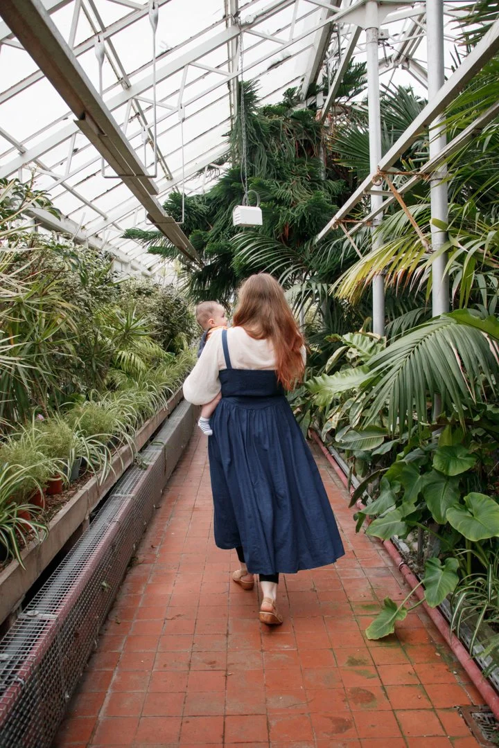 A woman with long red hair, wearing a blue dress, walking through a greenhouse with a young child in her arms, surrounded by lush green tropical plants.  Family Photography - Greenhouse Victoria Park, Stafford