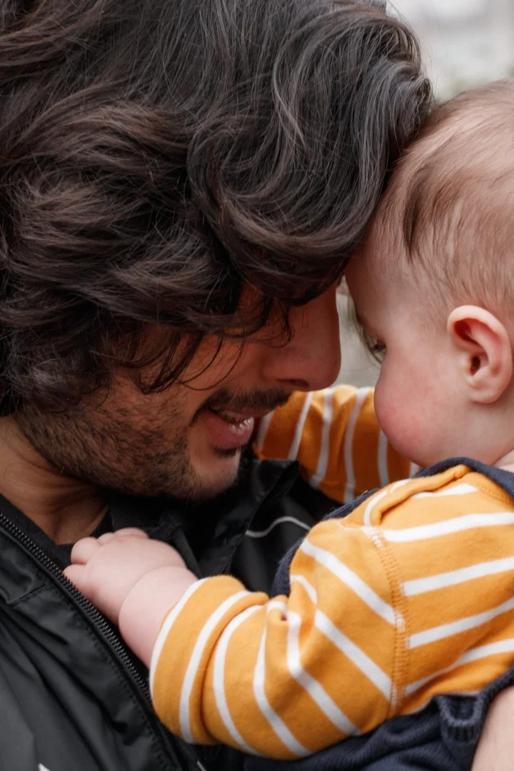 A man with dark, wavy hair and a beard is holding a young child with blonde hair, both touching foreheads and smiling. The child is wearing an orange and white striped shirt.  Family Photography