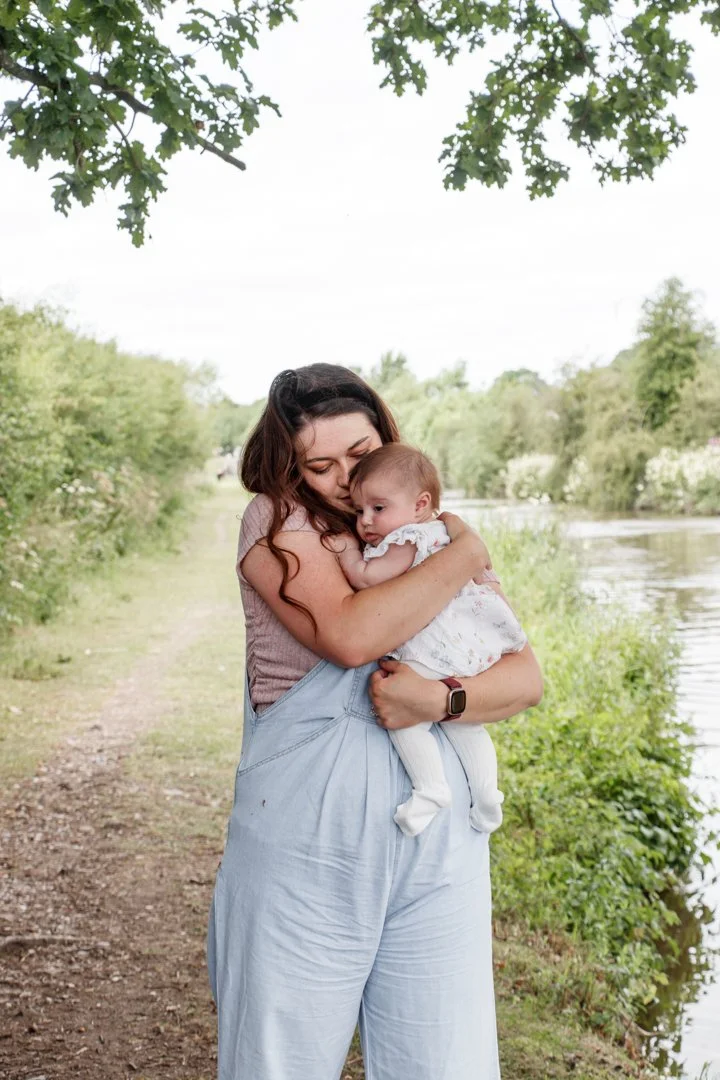 A woman holding a baby near a river or lake, surrounded by greenery and trees.  Family Photography - Aston Marina, Staffordshire