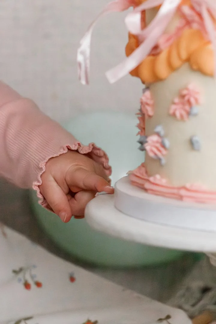Close-up of a child's hand touching a decorated cake with soft pastel colors and floral icing details. Cake smash