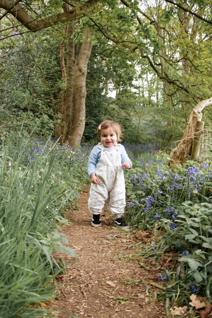 A young girl standing on a dirt path surrounded by green foliage and purple flowers in a wooded area.  Family Photography, Baby. Jackson's Coppice & Marsh