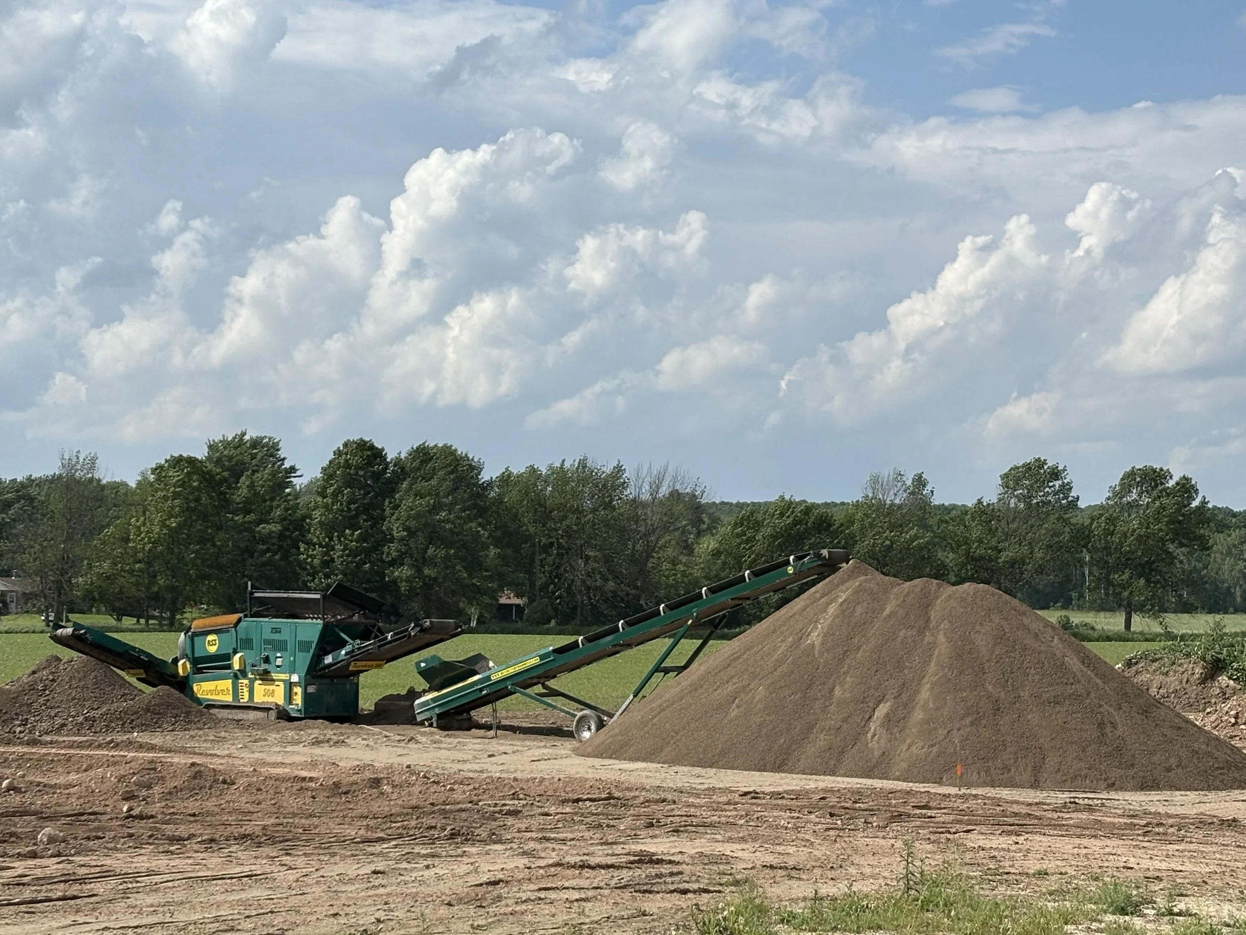 A construction site with heavy machinery and a pile of dirt under a partly cloudy sky, surrounded by trees.