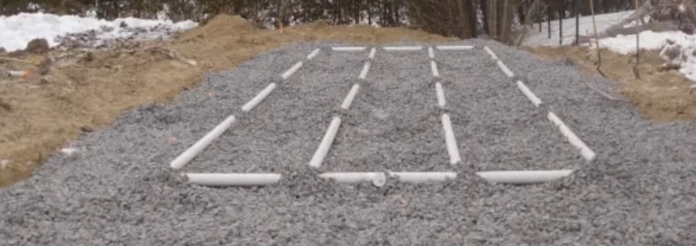 A construction site with gravel and white PVC pipes arranged in a grid pattern for a foundation or footing.