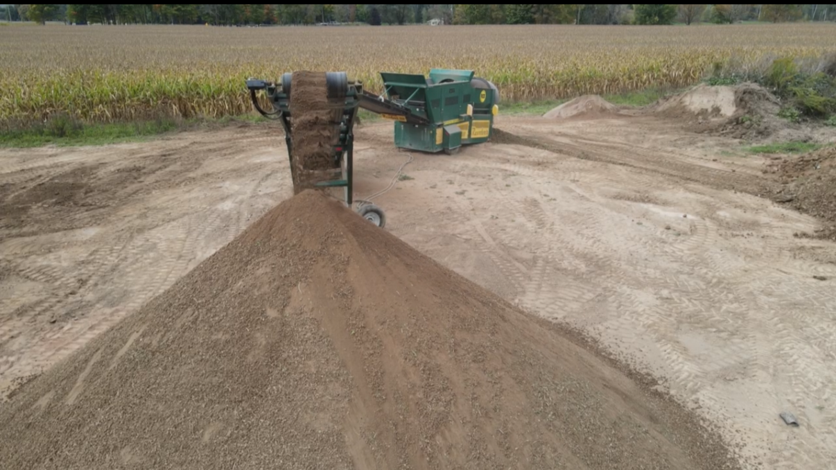 A dirt or soil screener and shredder machine in an open field with a background of crops, used for processing soil or compost on a farm.