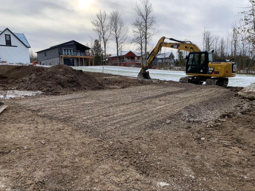 Construction site with a yellow excavator on a dirt surface, with residential houses and leafless trees in the background under an overcast sky.