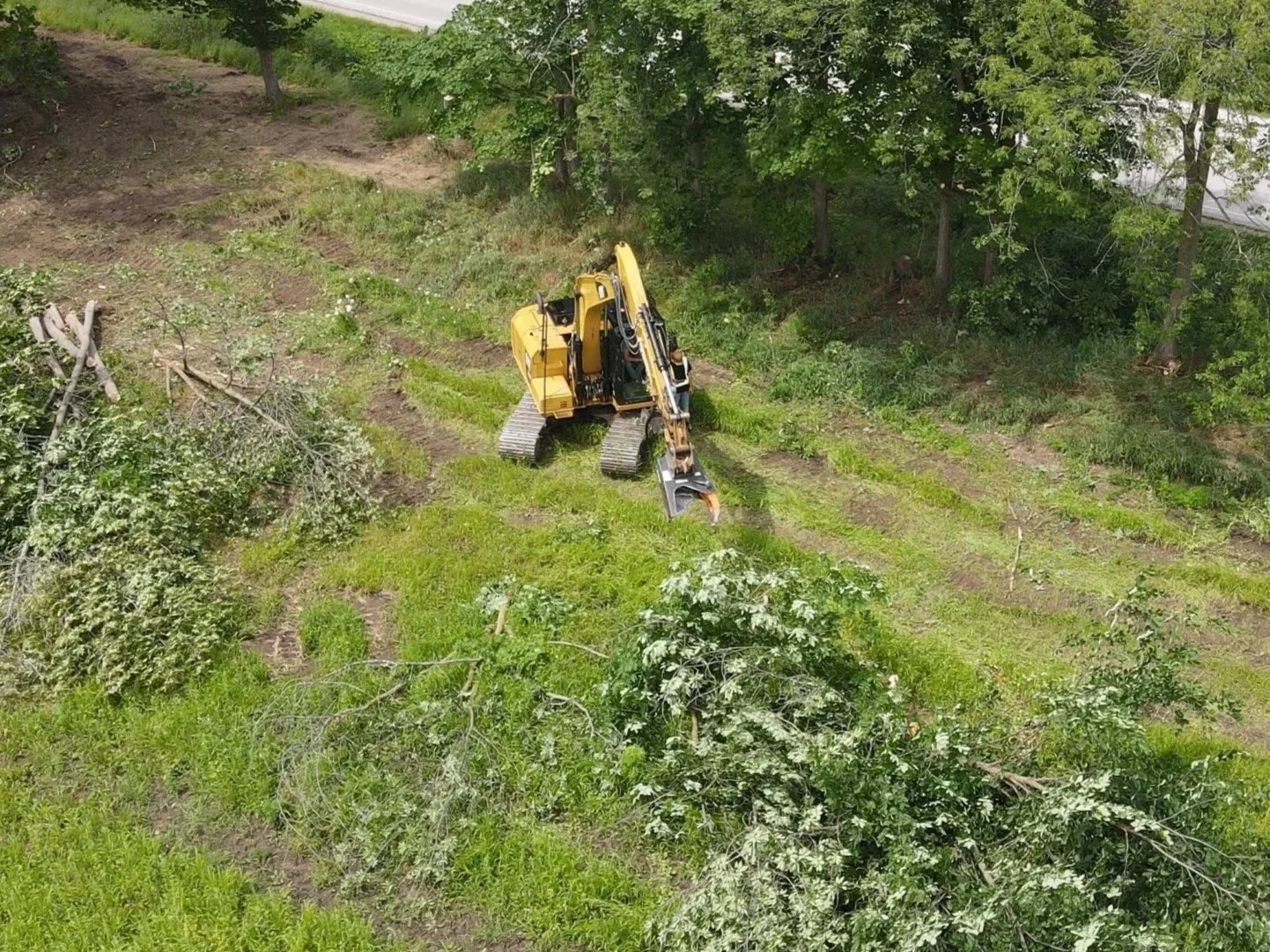 A yellow excavator clearing fallen tree branches and debris in a grassy area near a wooded border.