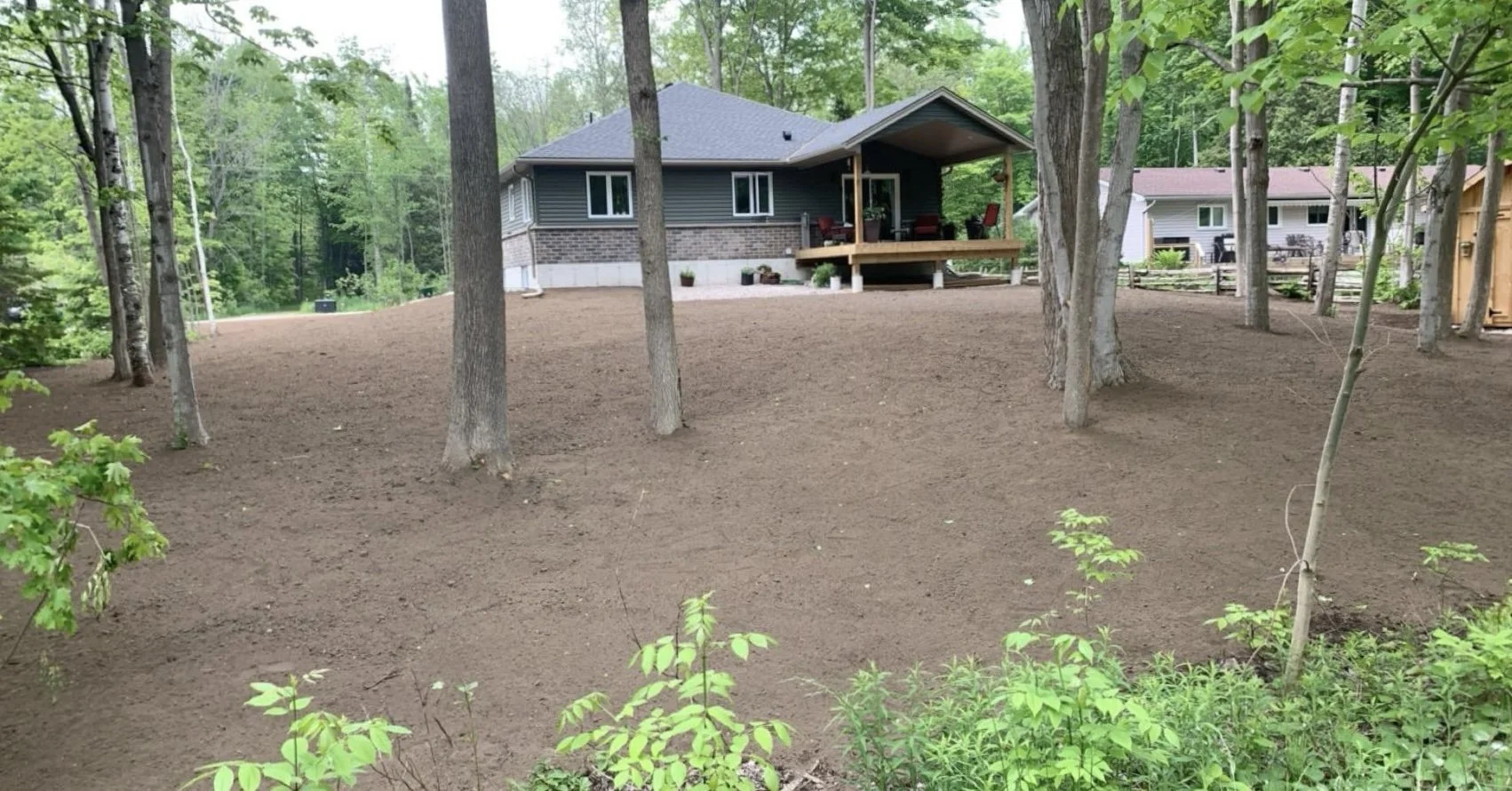 A backyard with freshly tilled soil, several trees, and a house with a porch in the background surrounded by green trees and other houses.