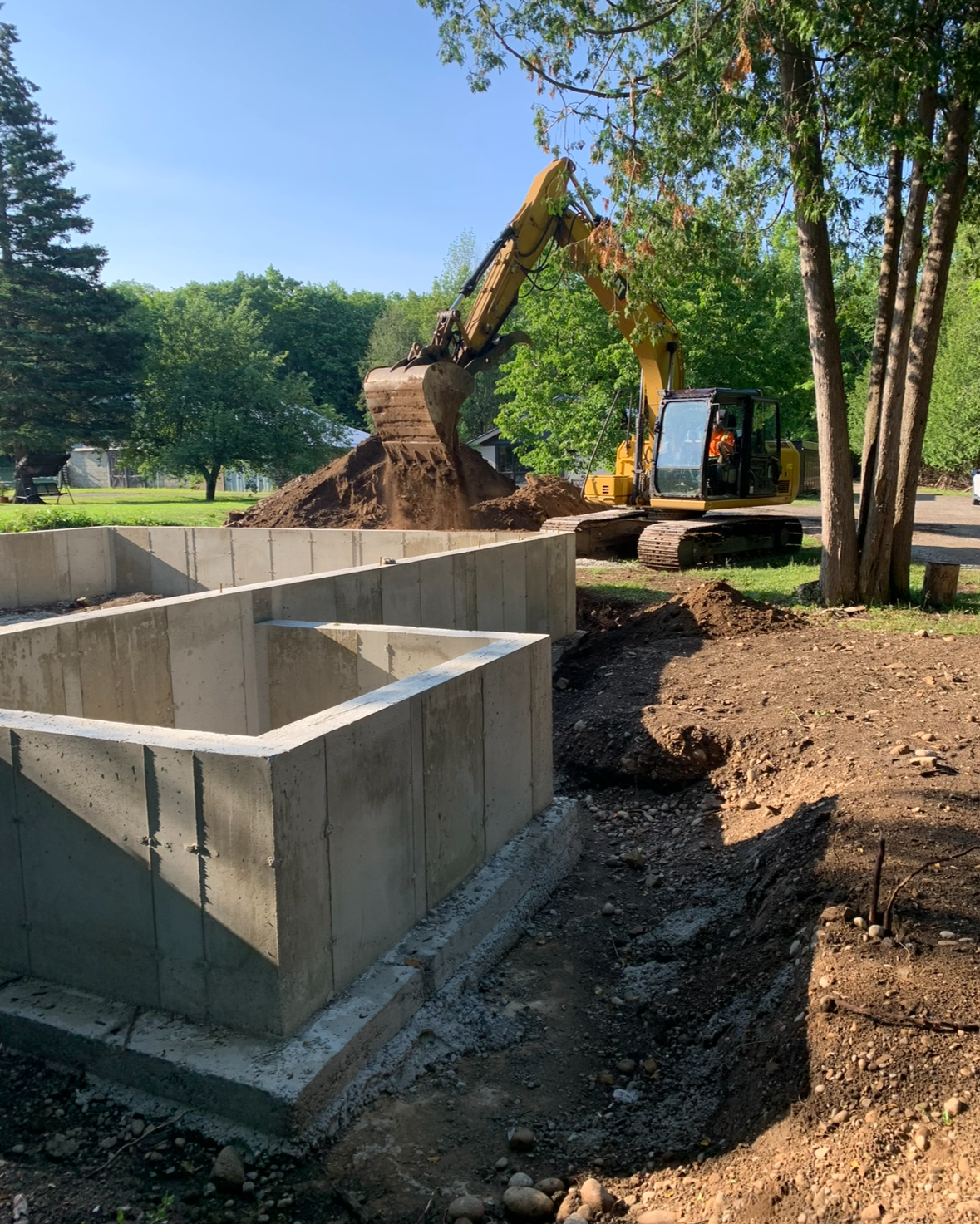 A yellow excavator is moving dirt into a concrete form for construction in a yard with green trees and a clear blue sky.