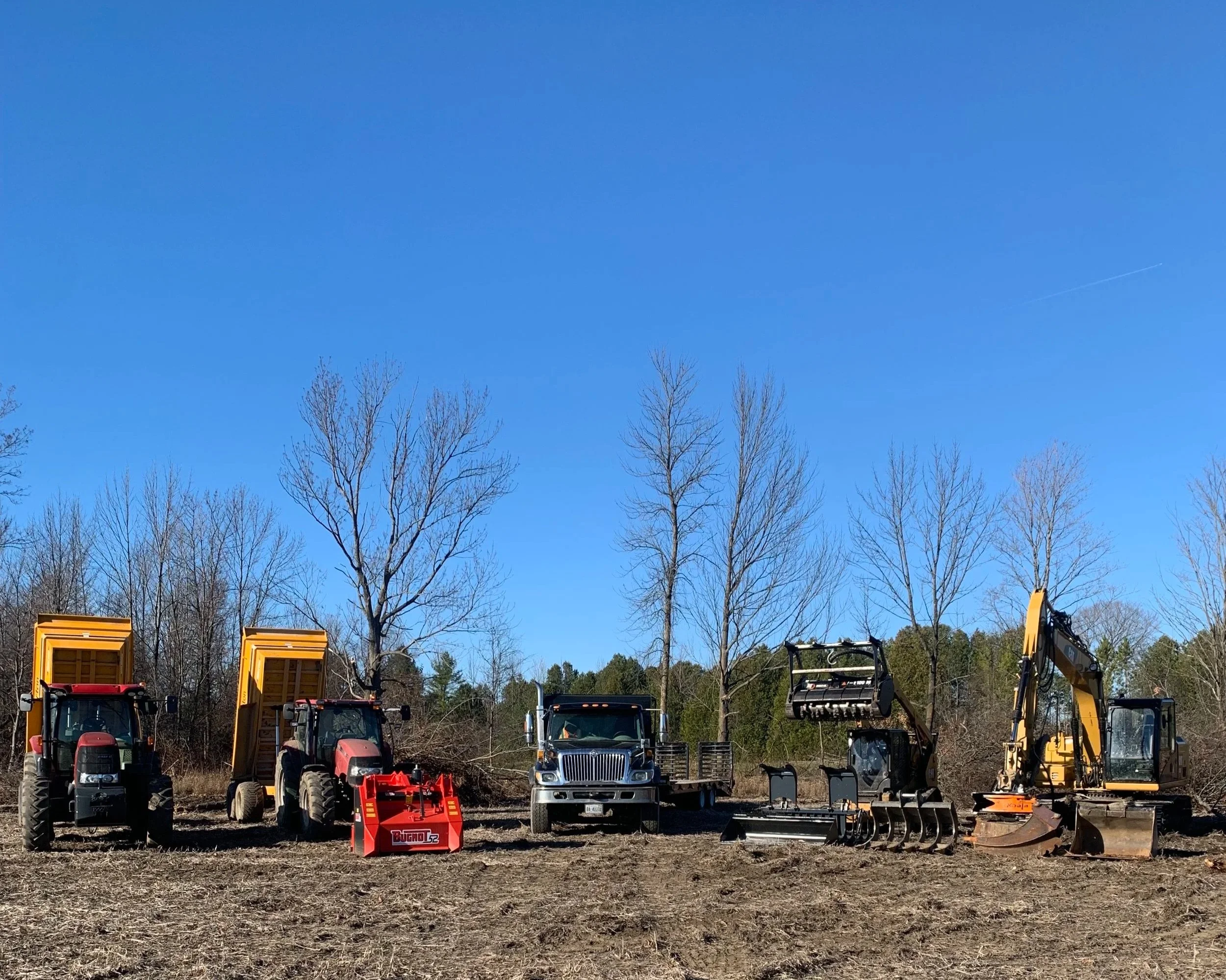 A line of various construction and farm equipment, including tractors and excavators, parked on a dirt lot with leafless trees and a clear blue sky in the background.