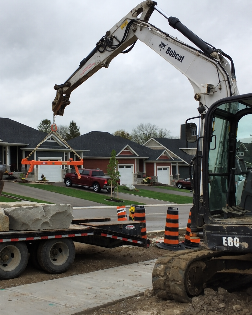 Construction site with a Bobcat E80 excavator lifting a large orange object, with orange and black striped traffic cones, a truck with large rocks, and a residential neighborhood with houses and parked cars in the background.