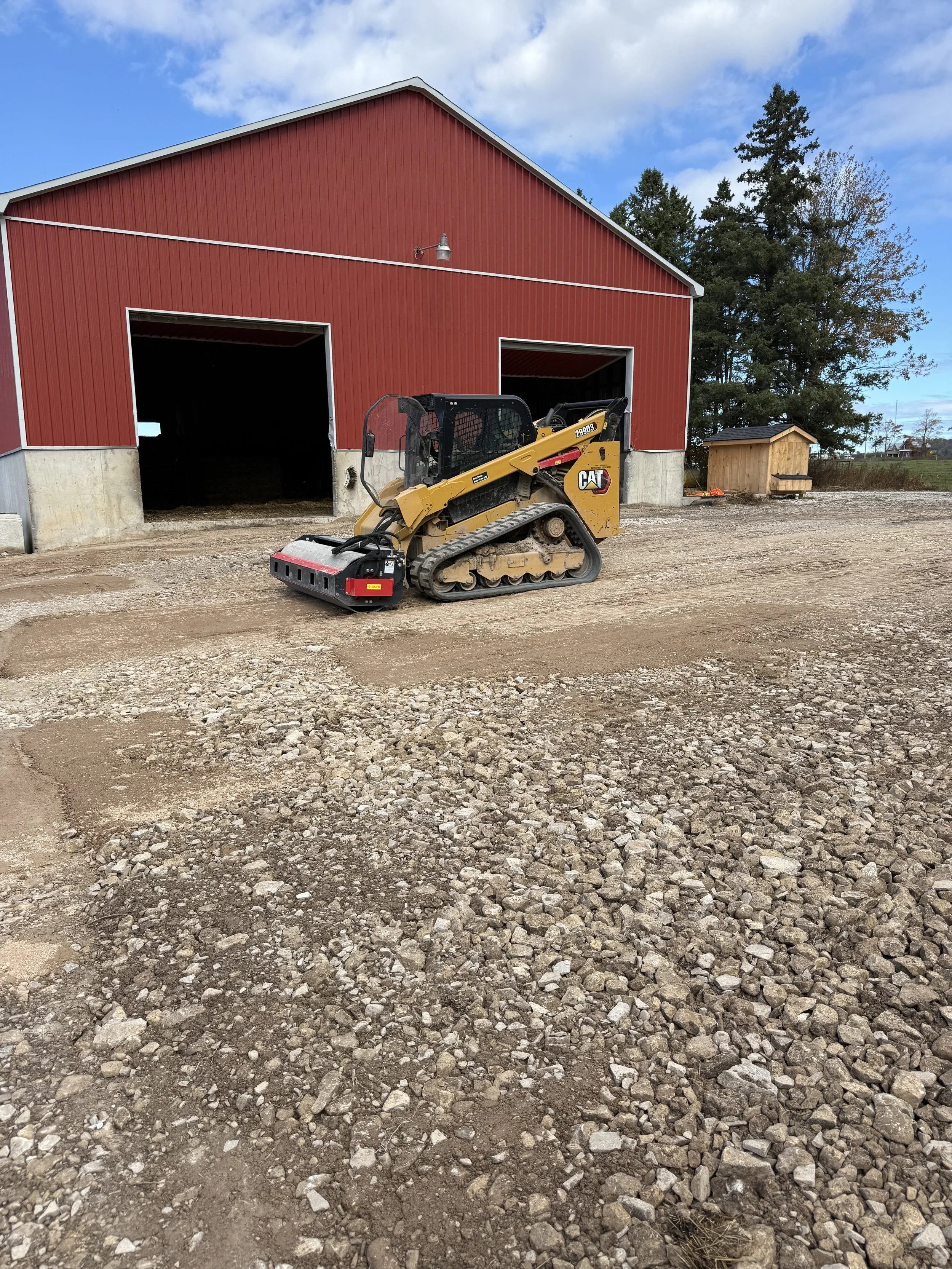 A small Caterpillar compact track loader parked on a dirt and gravel lot in front of a red barn with sliding doors. The sky is partly cloudy, and trees can be seen in the background.
