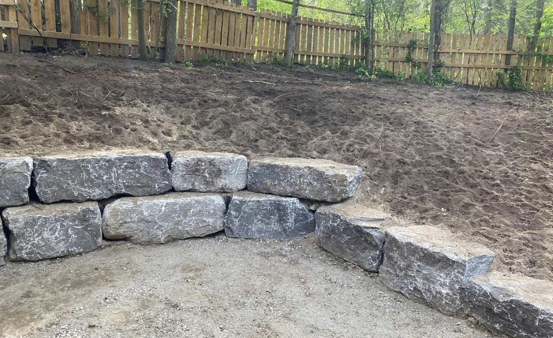 A retaining wall made of large gray stones in a backyard, with a dirt slope behind it and a wooden fence at the top.