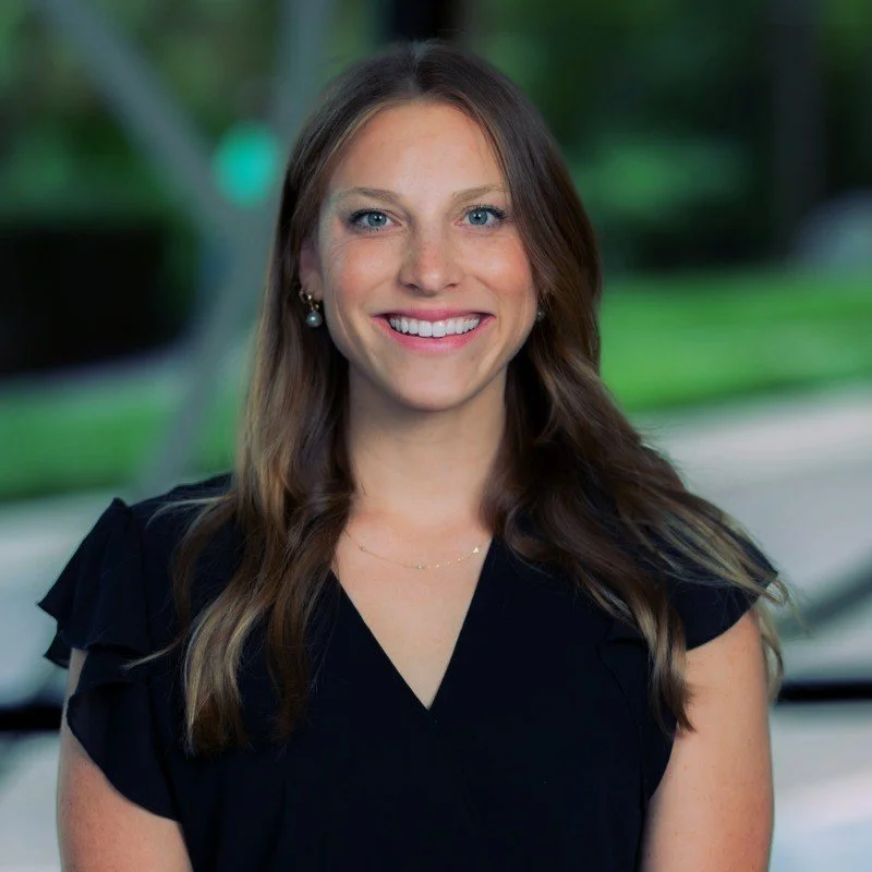 A woman with long brown hair wearing a black top, smiling outdoors with a blurred green background.