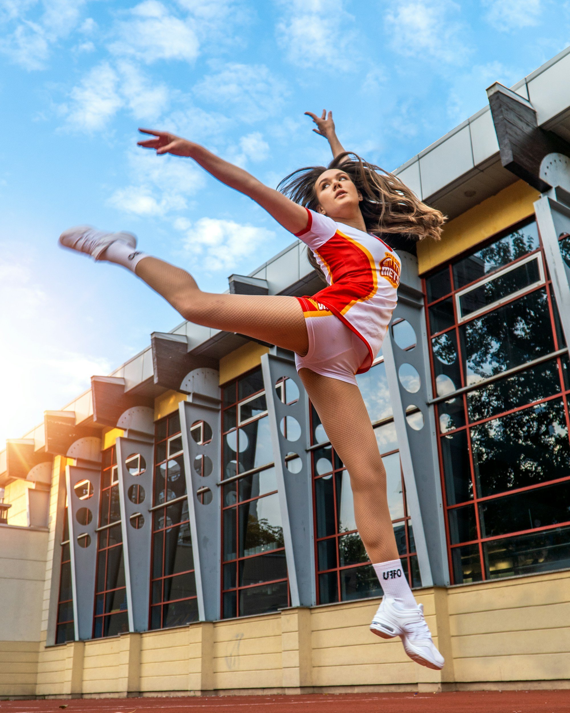 Young woman in cheerleading outfit performing a high jump outdoors during daytime, with modern building and blue sky in the background.