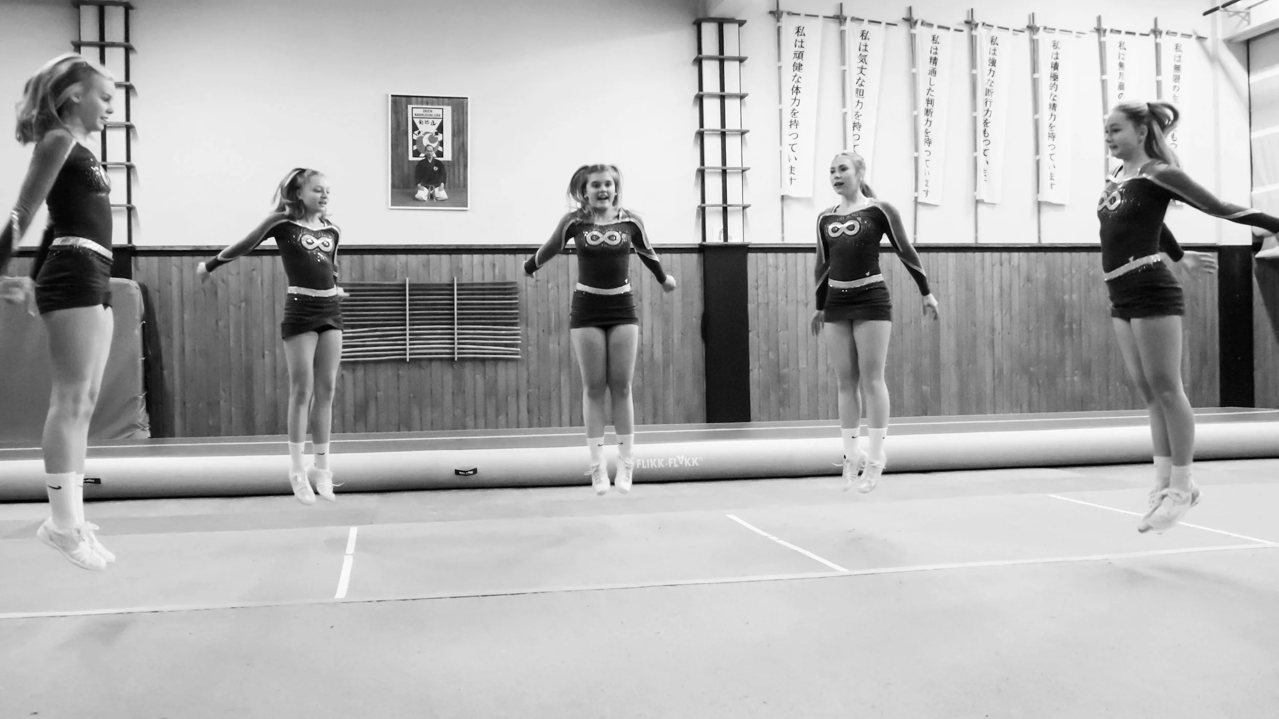 Five young girls in athletic uniforms jumping in unison on a gymnastics mat in a gymnasium.