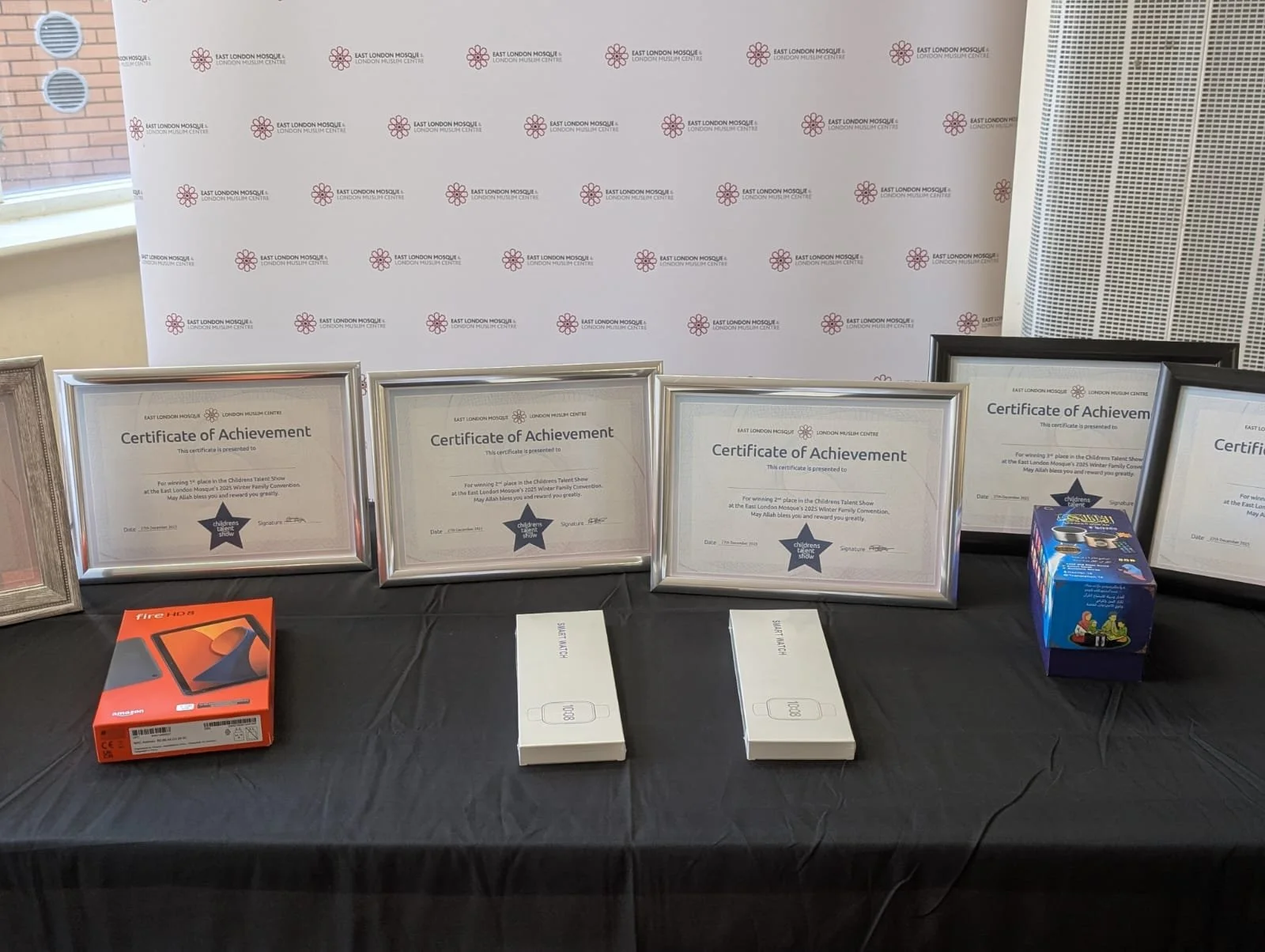 Table with four framed Certificates of Achievement, electronics boxes including an Amazon Fire HD 8 tablet, and other boxed items, set against a backdrop with a logo pattern for East London Mosque.
