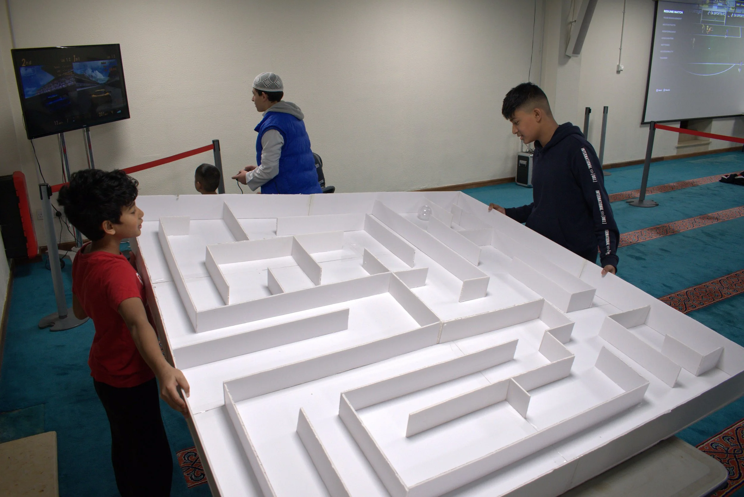 Three boys are gathered around a large white maze made from foam or cardboard, with one boy holding the edge, and two others observing. In the background, there are two screens displaying digital content, and two smaller children are present. The setting appears to be an indoor educational or recreational space.
