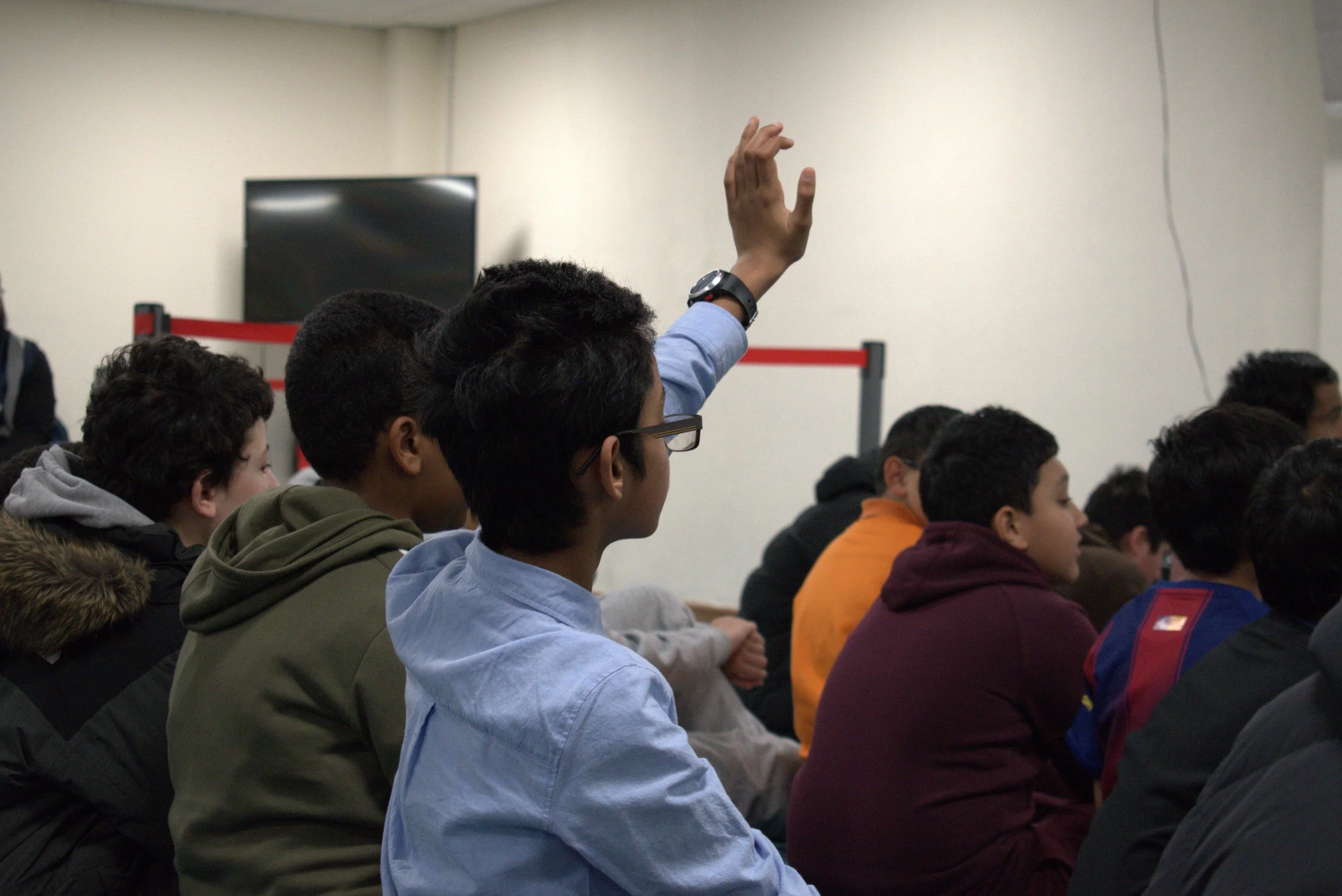 A classroom full of students sitting and attentively listening, with one student raising his hand.
