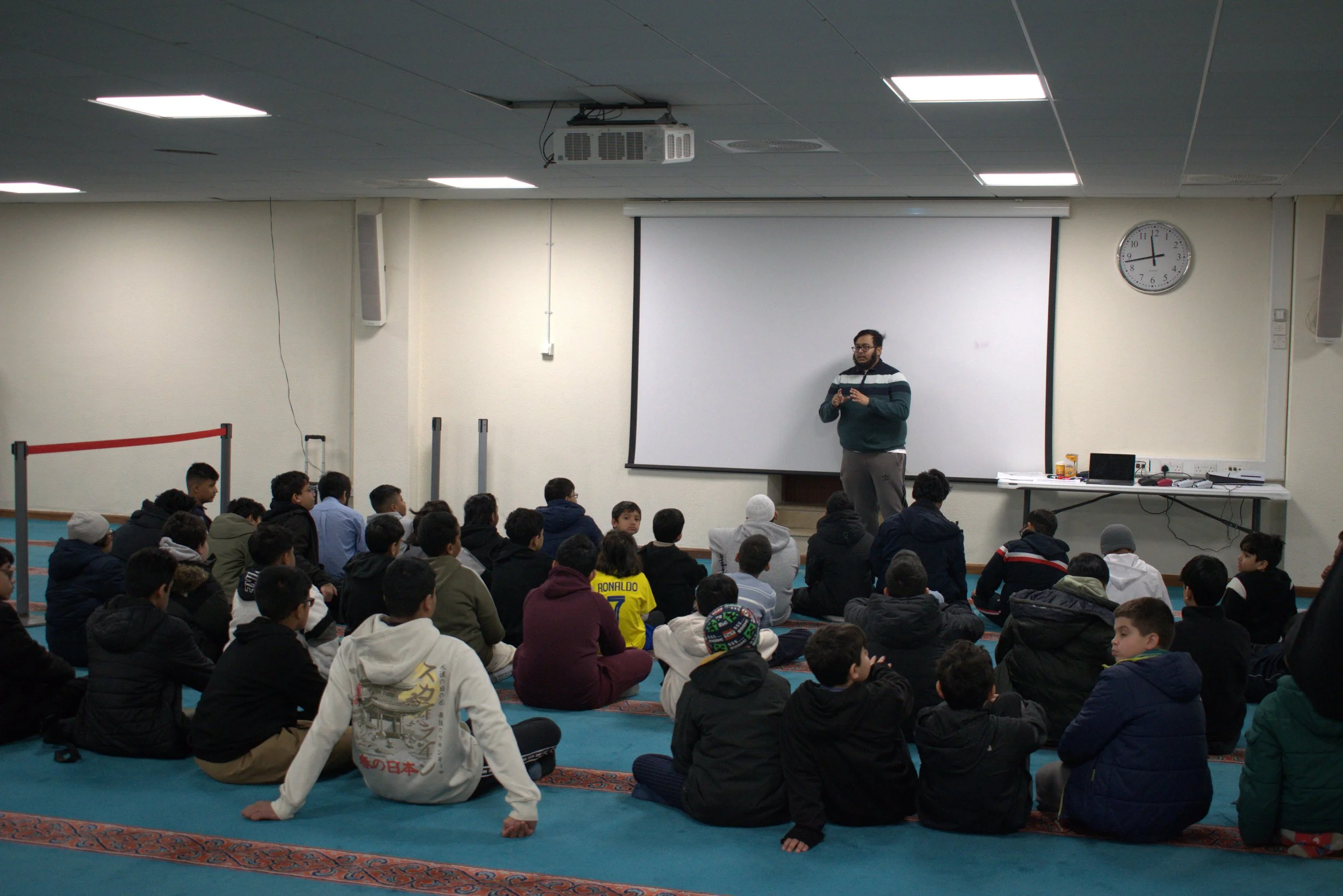 A group of children and teenagers are sitting on the floor facing a man who is standing in front of a whiteboard in a classroom or auditorium. The man appears to be giving a presentation or lecture.
