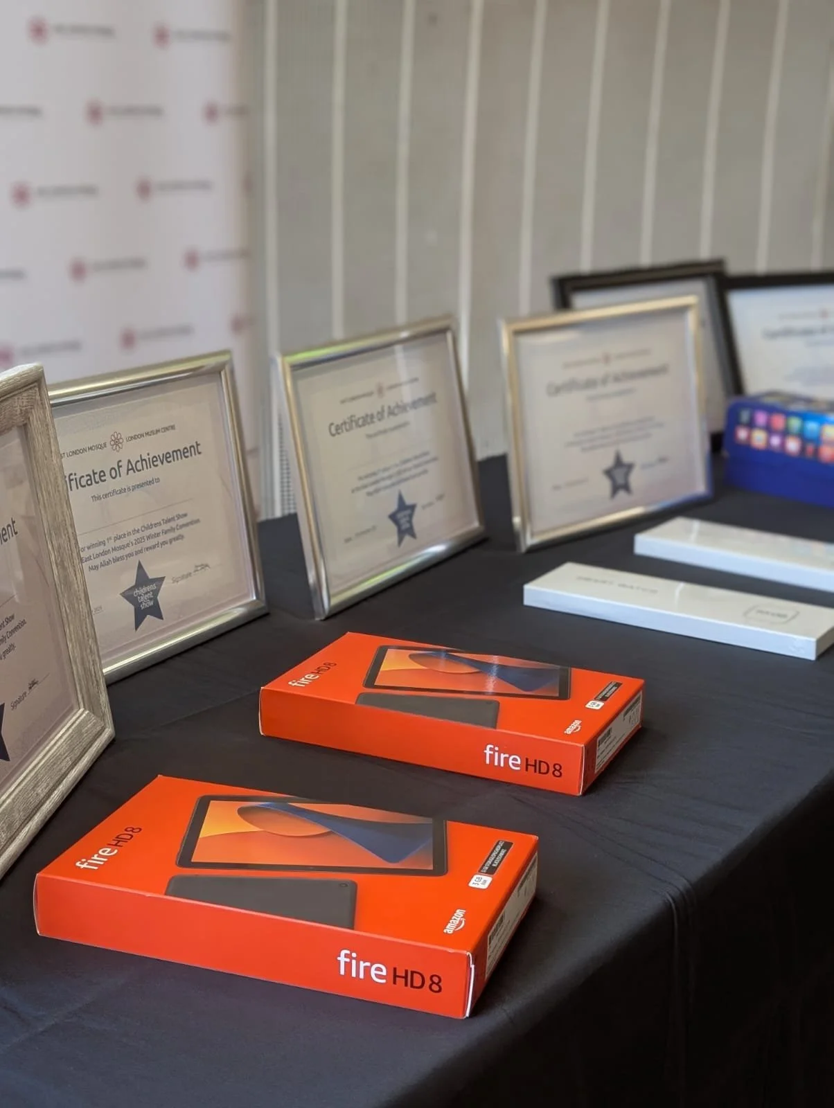 Table displaying two Amazon Fire HD 8 tablets in their orange boxes, with multiple framed certificates of achievement behind them on a black tablecloth.