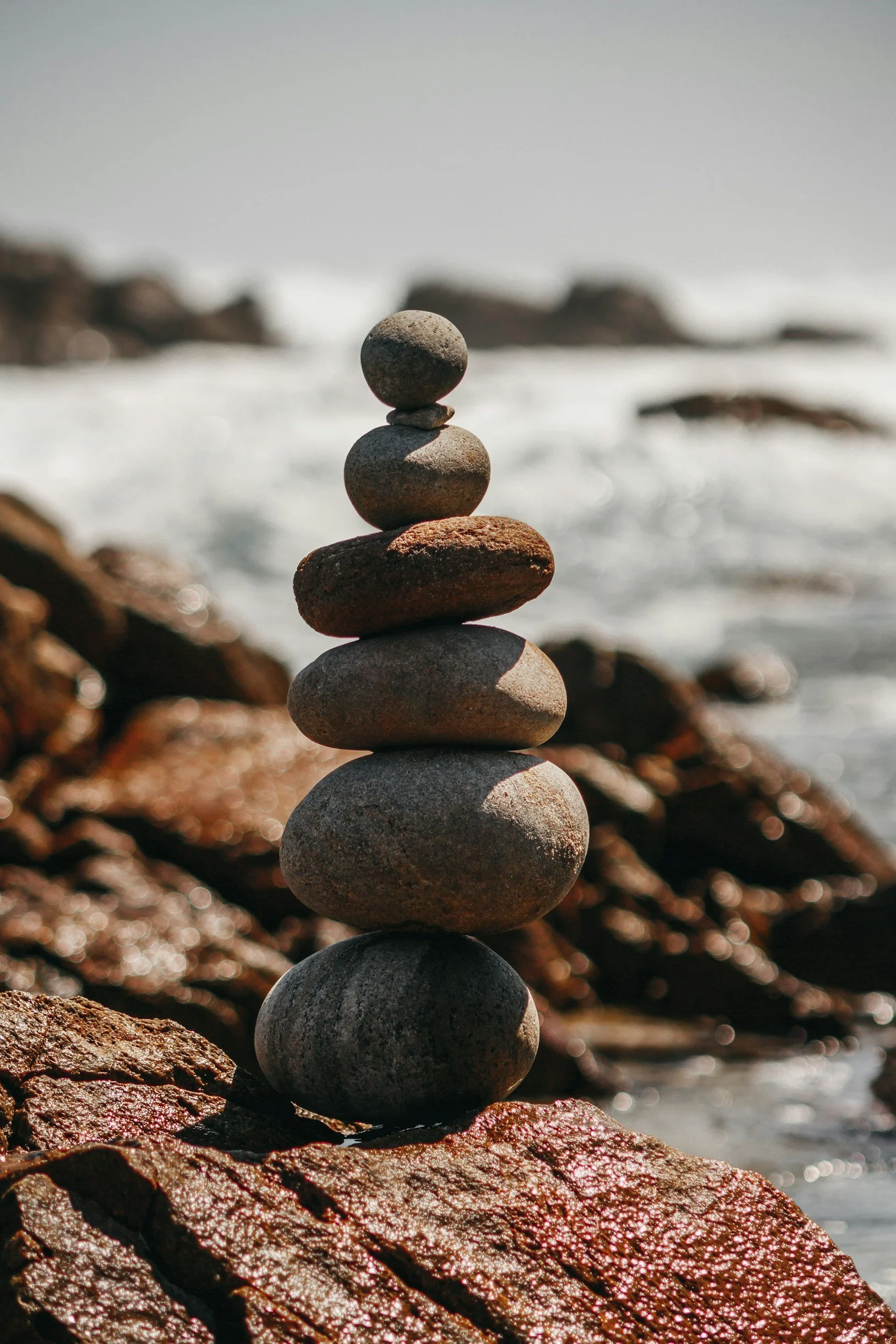 Stack of seven smooth stones balanced on a rocky beach with ocean waves in the background.