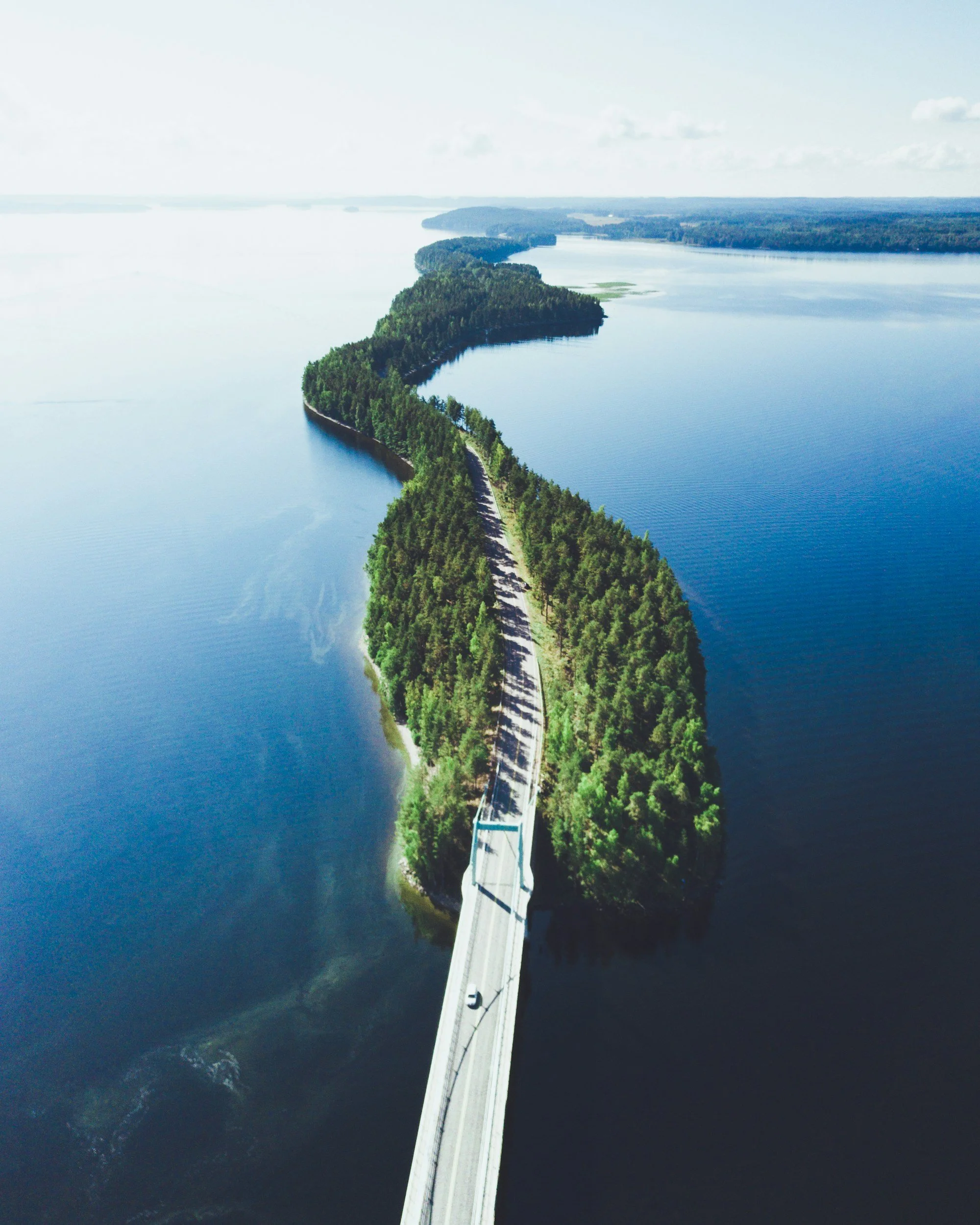 Ilmakuva pitkästä, kapeasta sillasta, joka ulottuu suuren vesistön yli ja jonka molemmin puolin kohoaa tiheä, vehreä metsä.