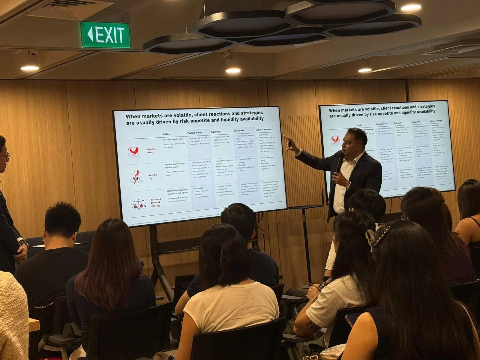 A man in a dark blazer and white shirt is giving a presentation in front of a room of seated audience members. He is pointing at a large screen displaying a presentation slide titled 'When markets are volatile, client reactions and strategies are usually driven by risk appetite and liquidity availability'. The room has wooden wall panels, a green exit sign, and ceiling decor.