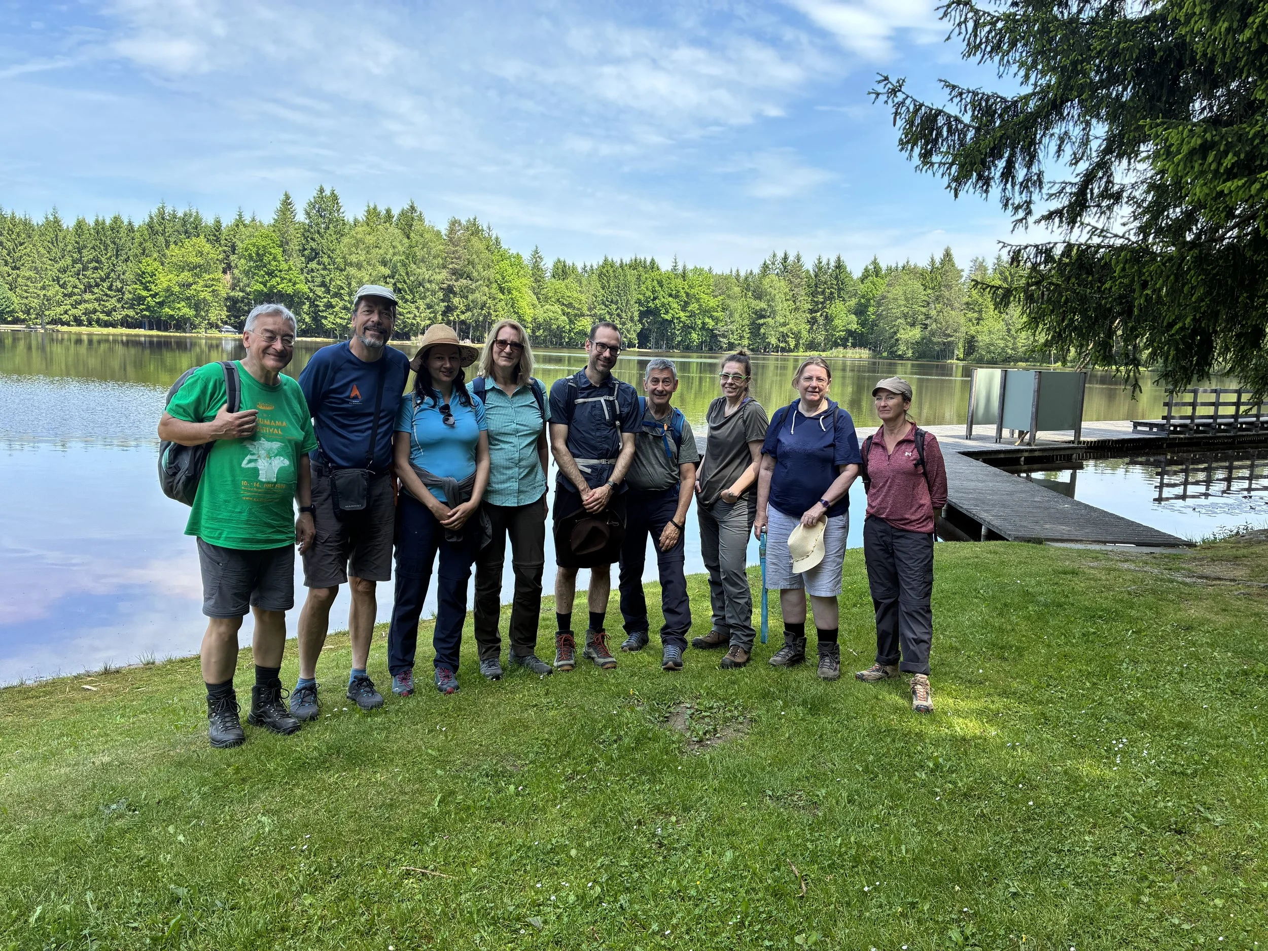Eine Gruppe von zehn Menschen steht am Ufer eines Sees mit Bäumen im Hintergrund. Es ist ein sonniger Tag, und sie scheinen eine Wanderung zu machen.