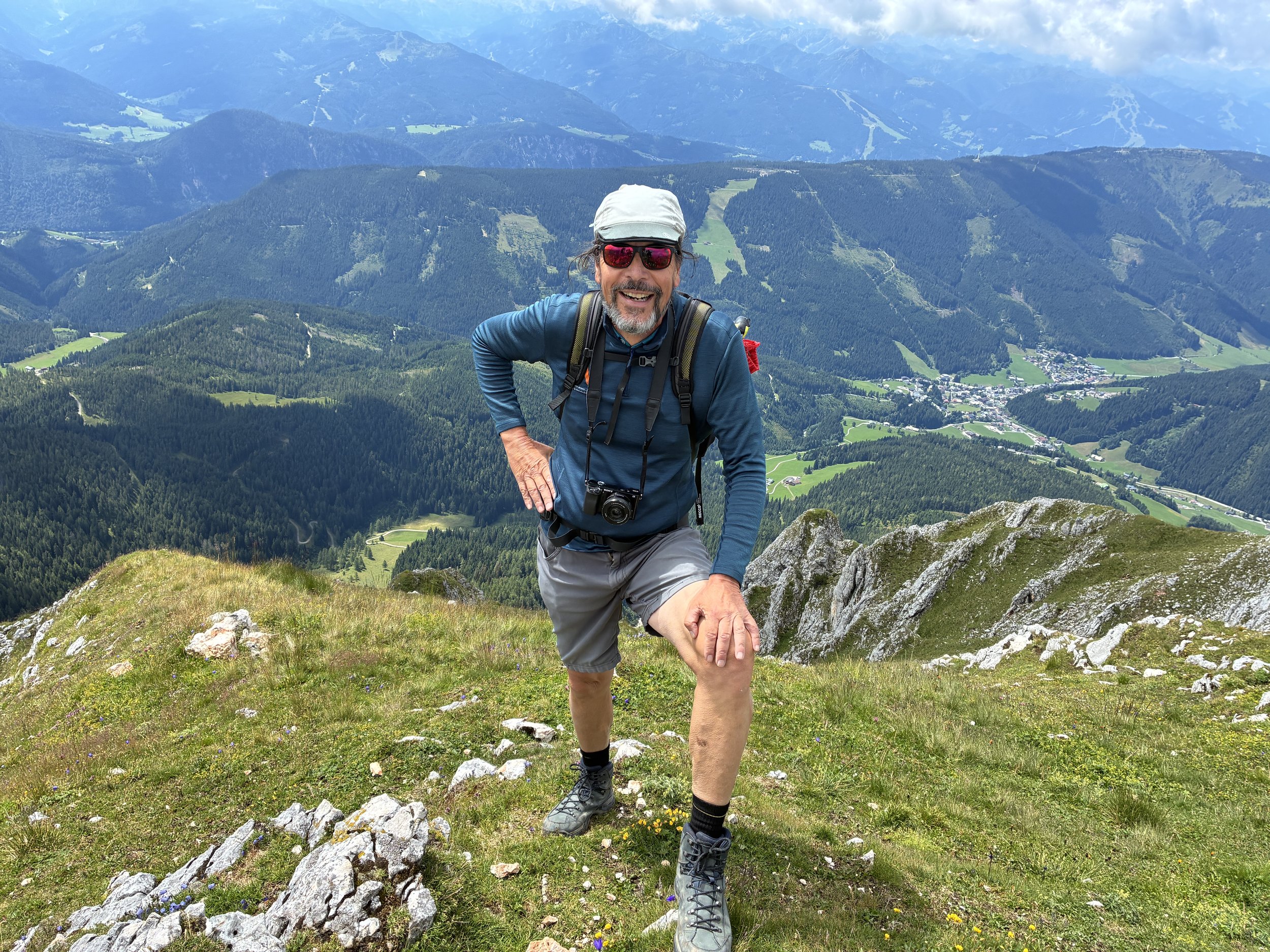 Ein Mann beim Wandern auf einem Berggipfel in den Alpen, trägt eine Mütze, Sonnenbrille, Rucksack und Kamera, mit einem Blick auf das Tal und die Berge im Hintergrund.