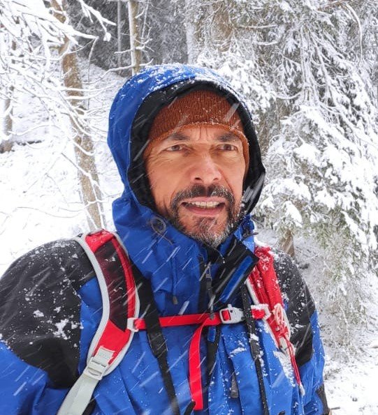 Mann beim Wandern im Winter mit Schneefall, Bäumen im Hintergrund, trägt blaue Jacke, rote Rucksack, Mütze.