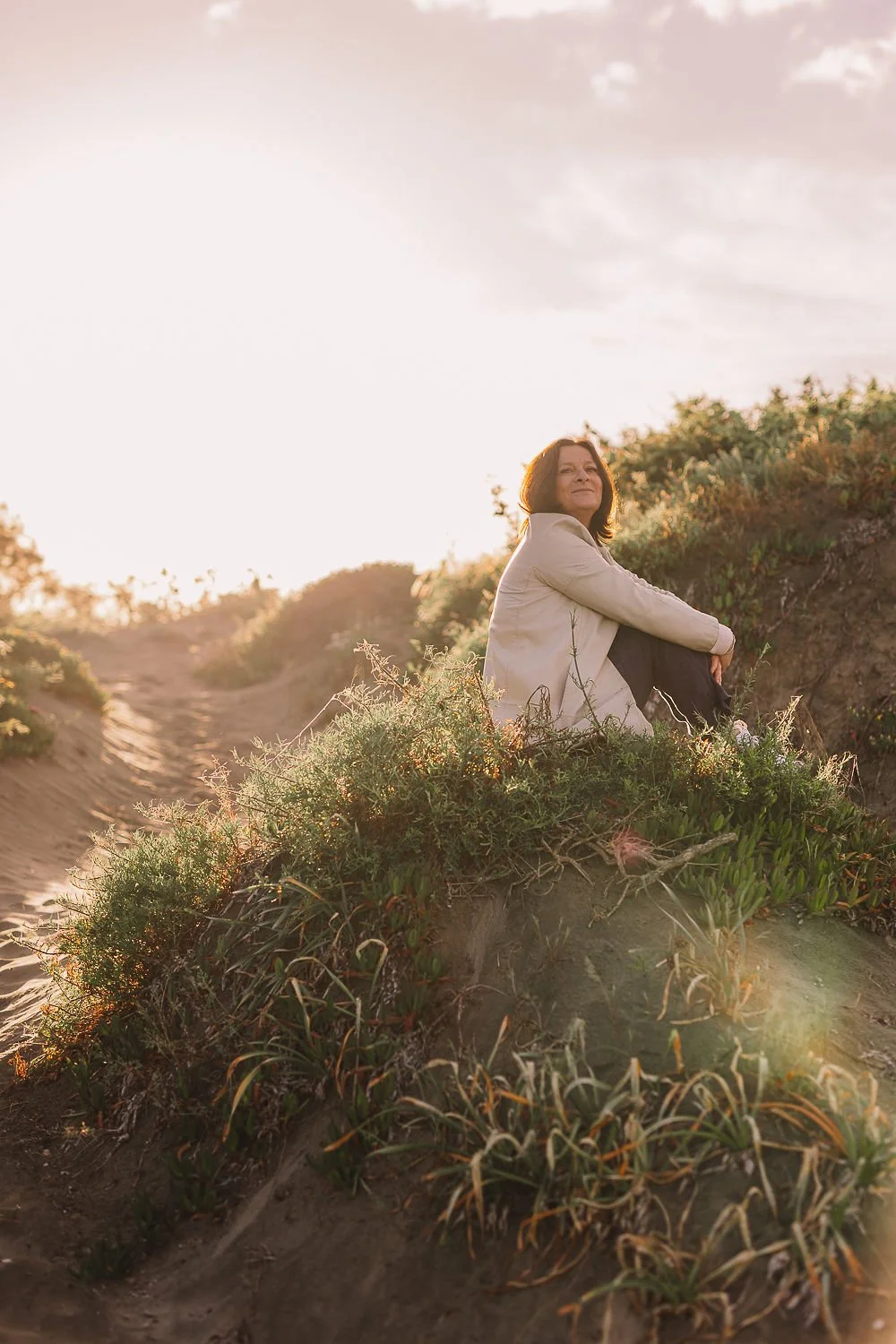 Una donna seduta su una duna di sabbia circondata da vegetazione, con il sole che tramonta all'orizzonte.