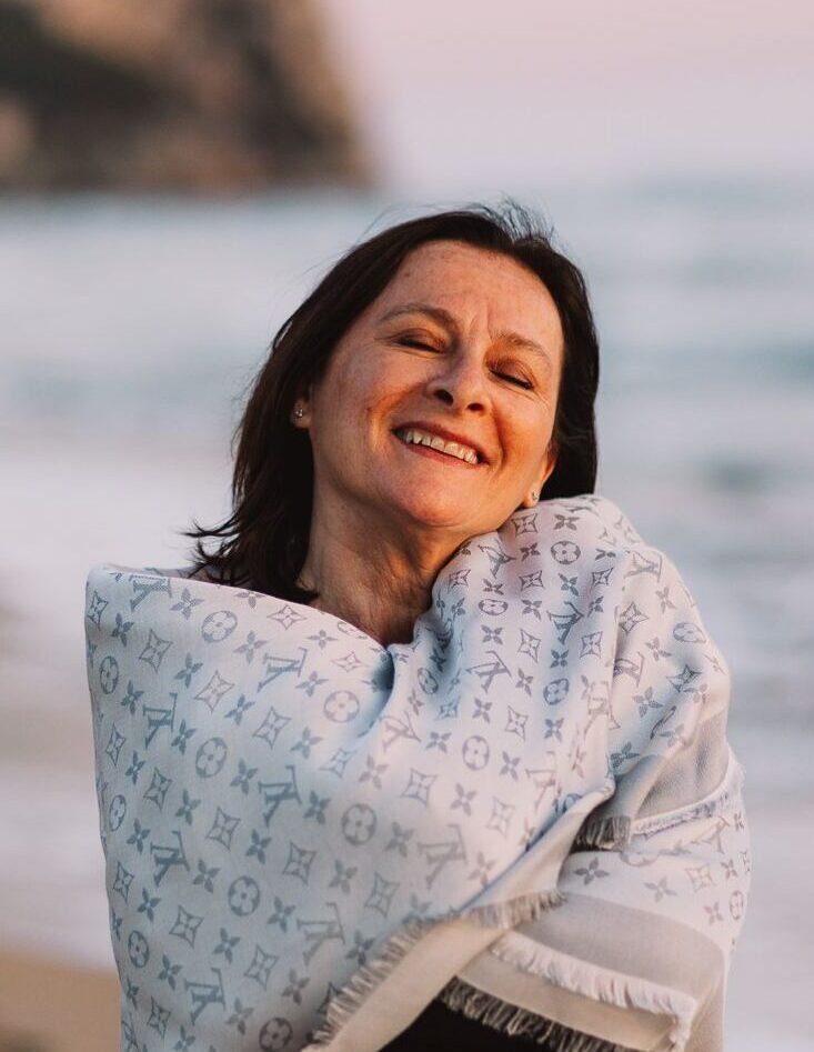 Donna sorridente che si trova sulla spiaggia al tramonto, avvolta in una coperta con motivo Louis Vuitton.
