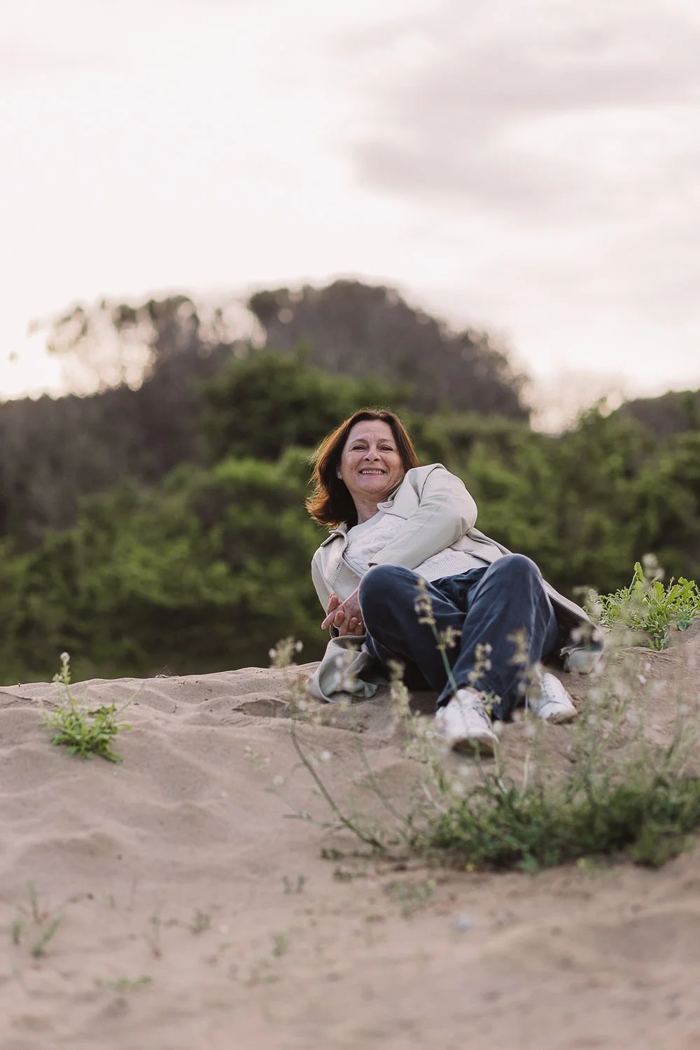 Una donna sorridente sdraiata sulla sabbia, con vegetazione verde e un cielo nuvoloso sullo sfondo.