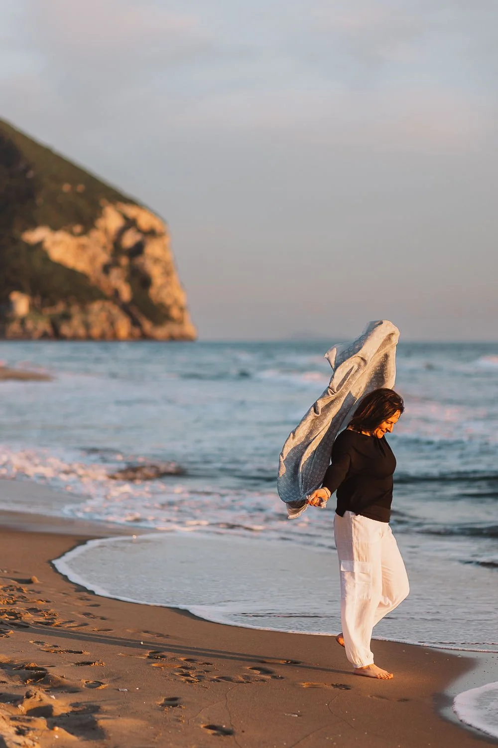 Una donna cammina sulla spiaggia al tramonto, con il mare e una scogliera sullo sfondo. È vestita con un maglione nero e pantaloni chiari, tiene un telo o uno scialle dietro le spalle.