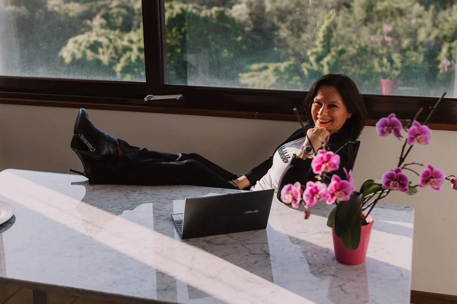 Una donna sorridente si rilassa con i piedi sul tavolo di marmo, seduta di fronte a una finestra che mostra un paesaggio verde. Sul tavolo c'è un laptop ASUS e un vaso con orchidee rosa.