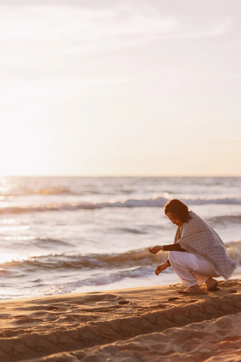 Una donna inginocchiata sulla spiaggia al tramonto, che si prende cura di qualcosa vicino al mare.