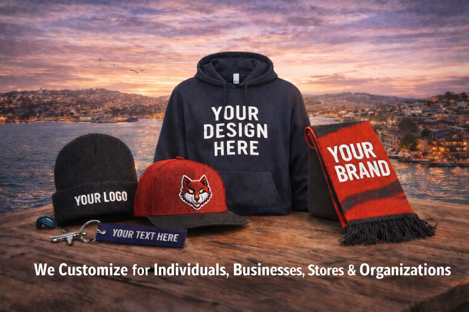 Customizable merchandise displayed on a wooden surface with a scenic water and cityscape sunset background, including a black hoodie with 'YOUR DESIGN HERE', a black beanie with 'YOUR LOGO', a red and black cap with a wolf logo, a red and black scarf with 'YOUR BRAND', and a blue keychain with a gun shape and a 'YOUR TEXT HERE' tag. Text at the bottom states, "We Customize for Individuals, Businesses, Stores & Organizations."
