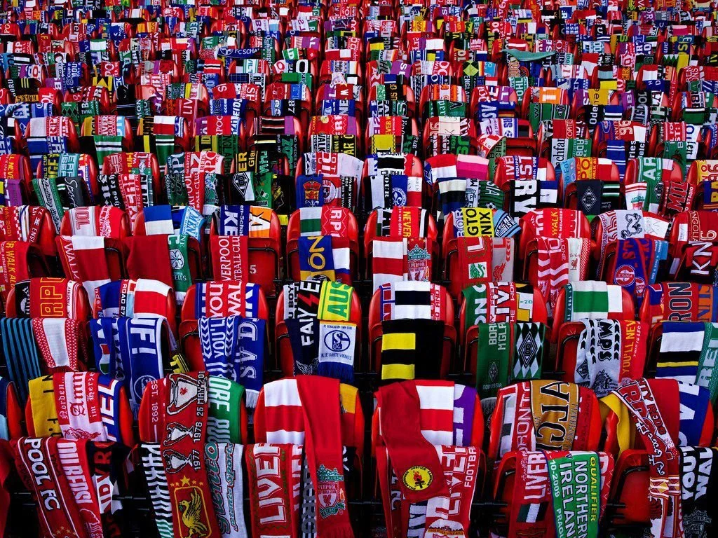 A large collection of red stadium seats covered with various team scarves, each scarf displaying team colors, logos, and names, creating a vibrant mosaic of soccer memorabilia.